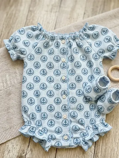 Hellblauer Baby-Romper und passende Schühchen aus Musselin-Stoff mit maritimem Anker-Muster und Wellen-Struktur, dekoriert mit Holzknöpfen auf einer Strickunterlage.