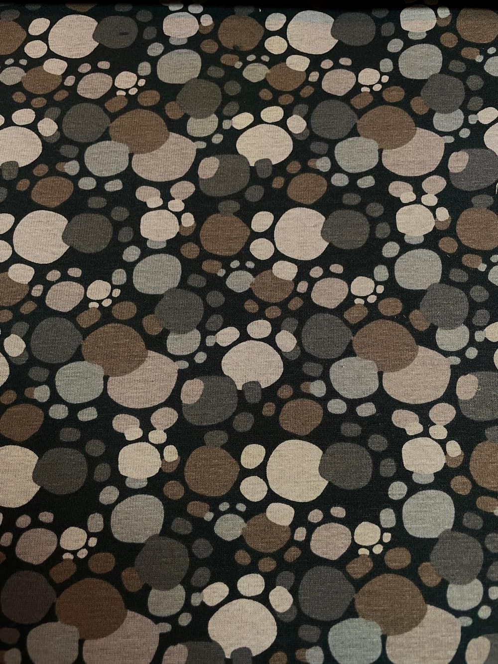 Dunkler Alpenfleece-Stoff mit einem Muster aus abstrakten Pfotenabdrücken und Punkten in verschiedenen Erdtönen wie Braun, Taupe, Beige und Grau auf schwarzem Hintergrund.