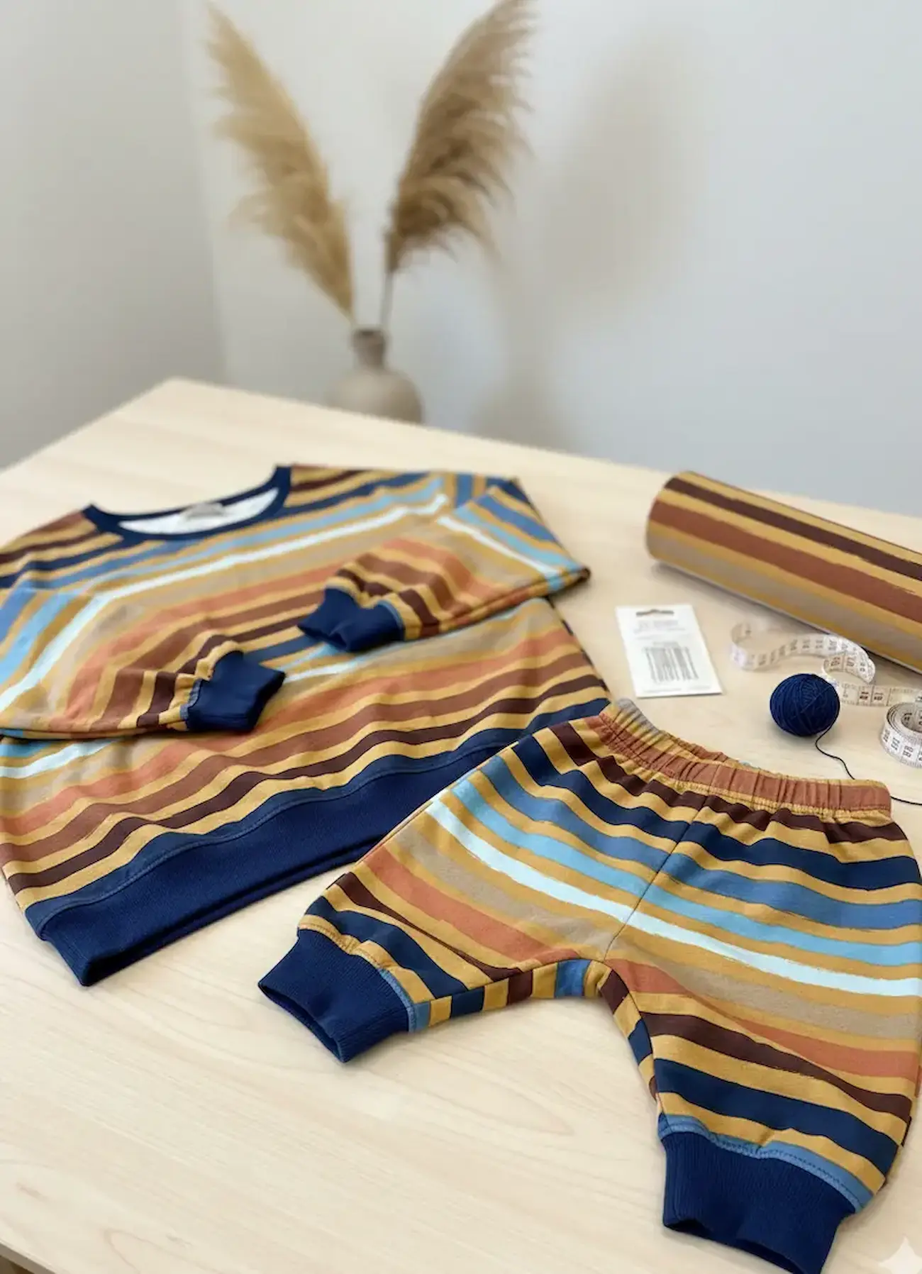 Bunt gestreifter French Terry Stoff als Meterware auf der Rolle neben einem fertig genähten Baby-Sweatshirt und einer Hose auf einem hellen Holztisch.