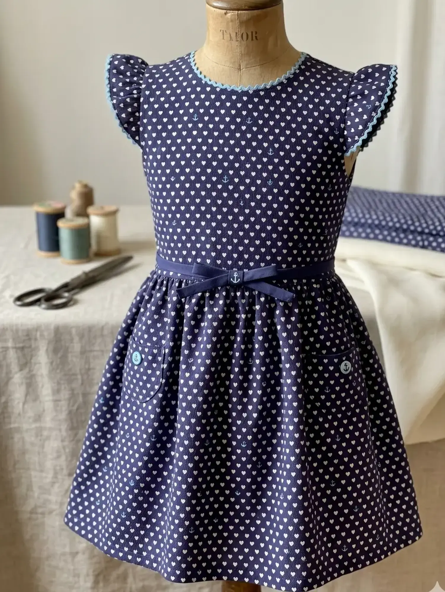 Dunkelblaues Kinderkleid mit Flügelärmeln aus Mors Crepe Baumwolle, verziert mit weißem Herzmuster, Zackenlitze am Ausschnitt und einer Schleife mit Anker-Motiv.