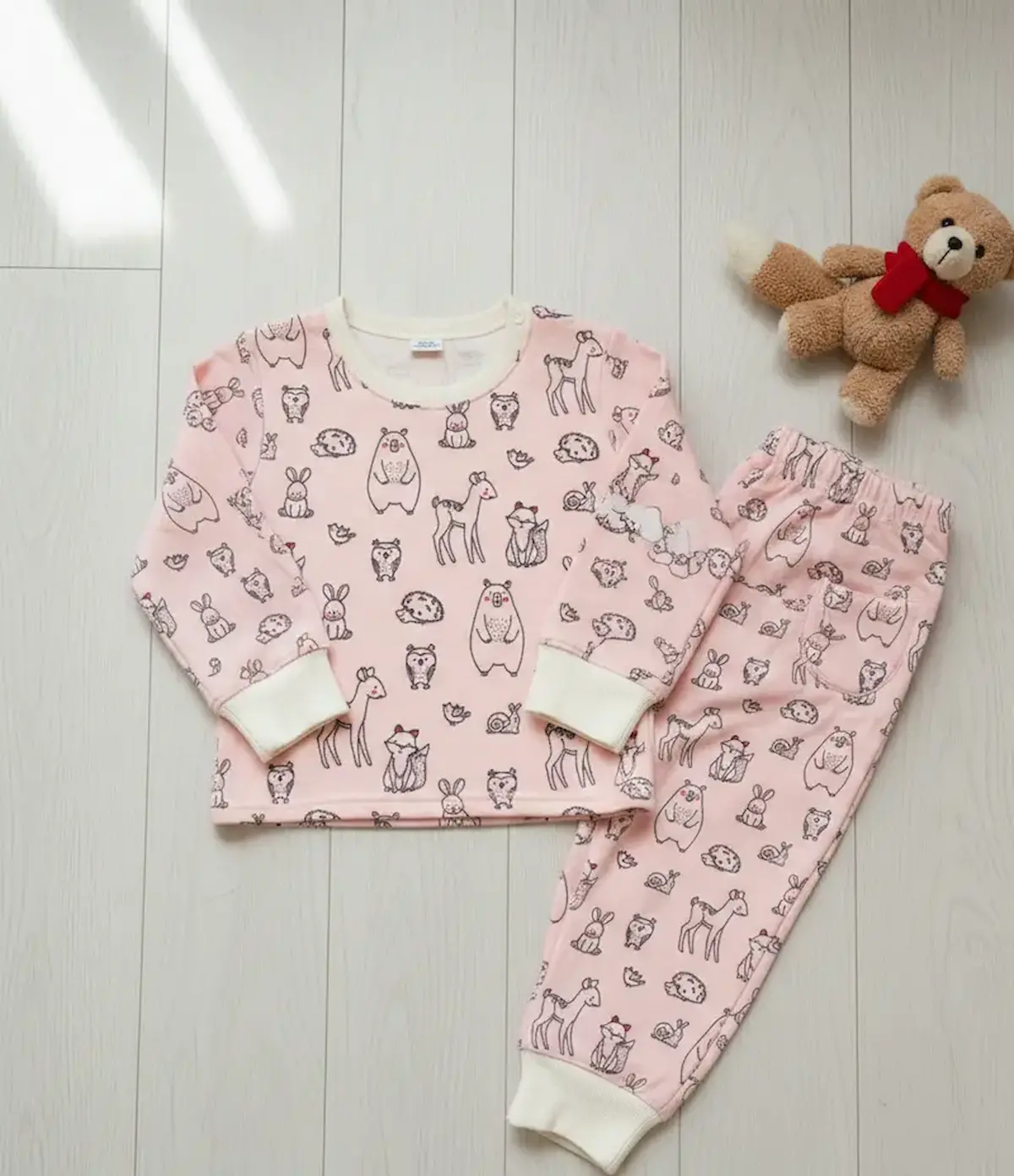 Zweiteiliges Kinder-Set aus rosa French Terry mit Waldtier-Motiven, bestehend aus Langarmshirt und Hose, flach liegend auf hellem Boden neben einem Teddybär.
