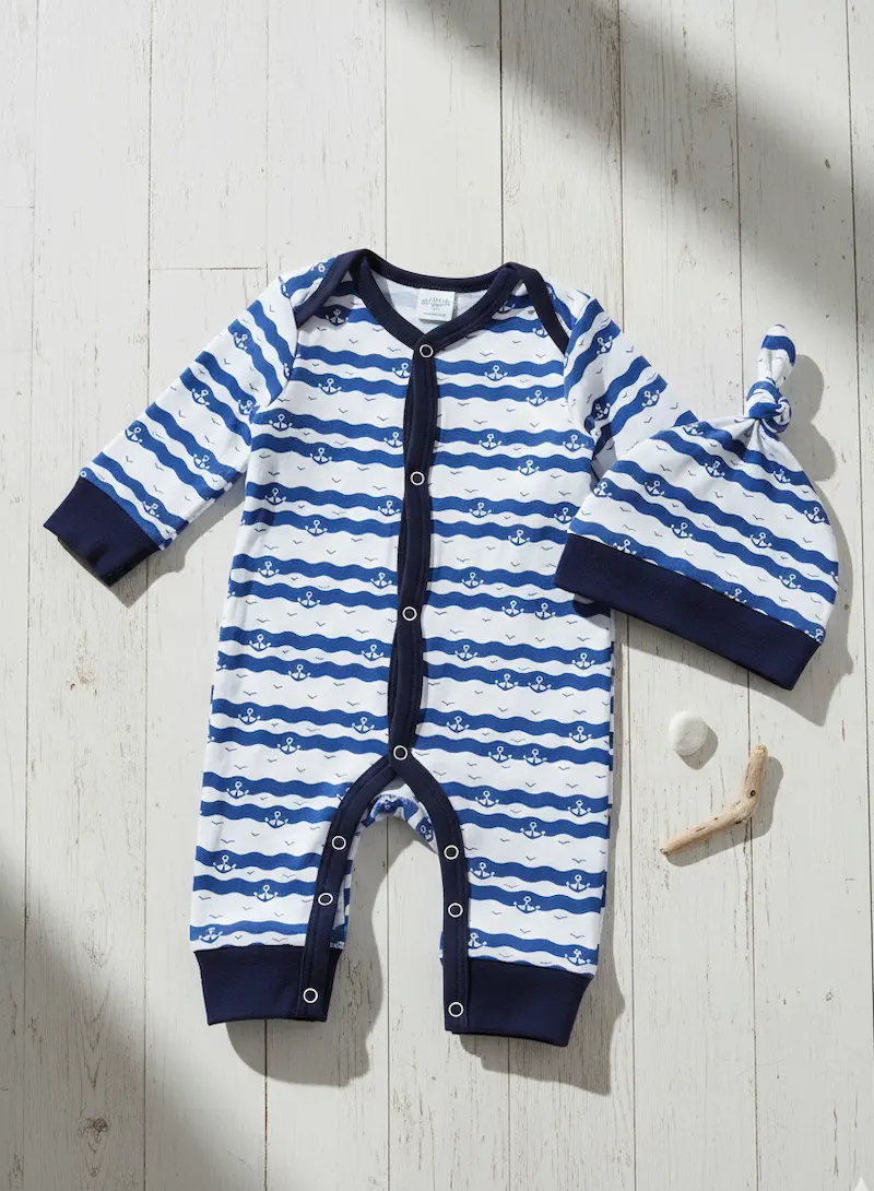 Weiß-blau gestreifter Baumwolljersey mit Wellen und kleinen Ankern, genäht als Baby-Strampler und Knotenmütze auf hellem Holzboden.