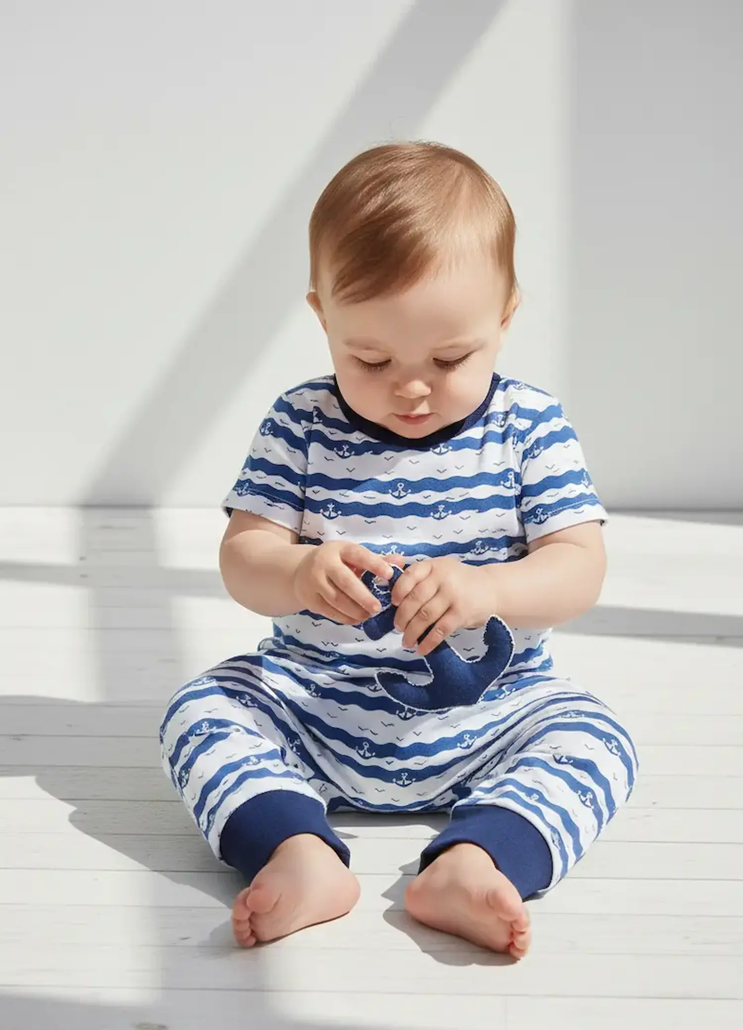 Ein Baby sitzt in einem kurzärmeligen Strampler aus blau-weißem Baumwolljersey mit Wellen- und Ankermotiv auf hellem Boden und spielt mit einem Stoffanker.