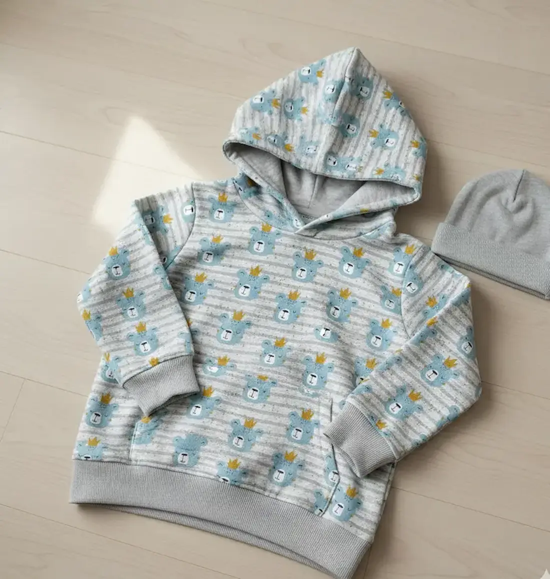 French Terry Stoff in Grau-Melange mit hellblauen Bären-Köpfen und goldenen Kronen, flach liegend als Hoodie mit Mütze.