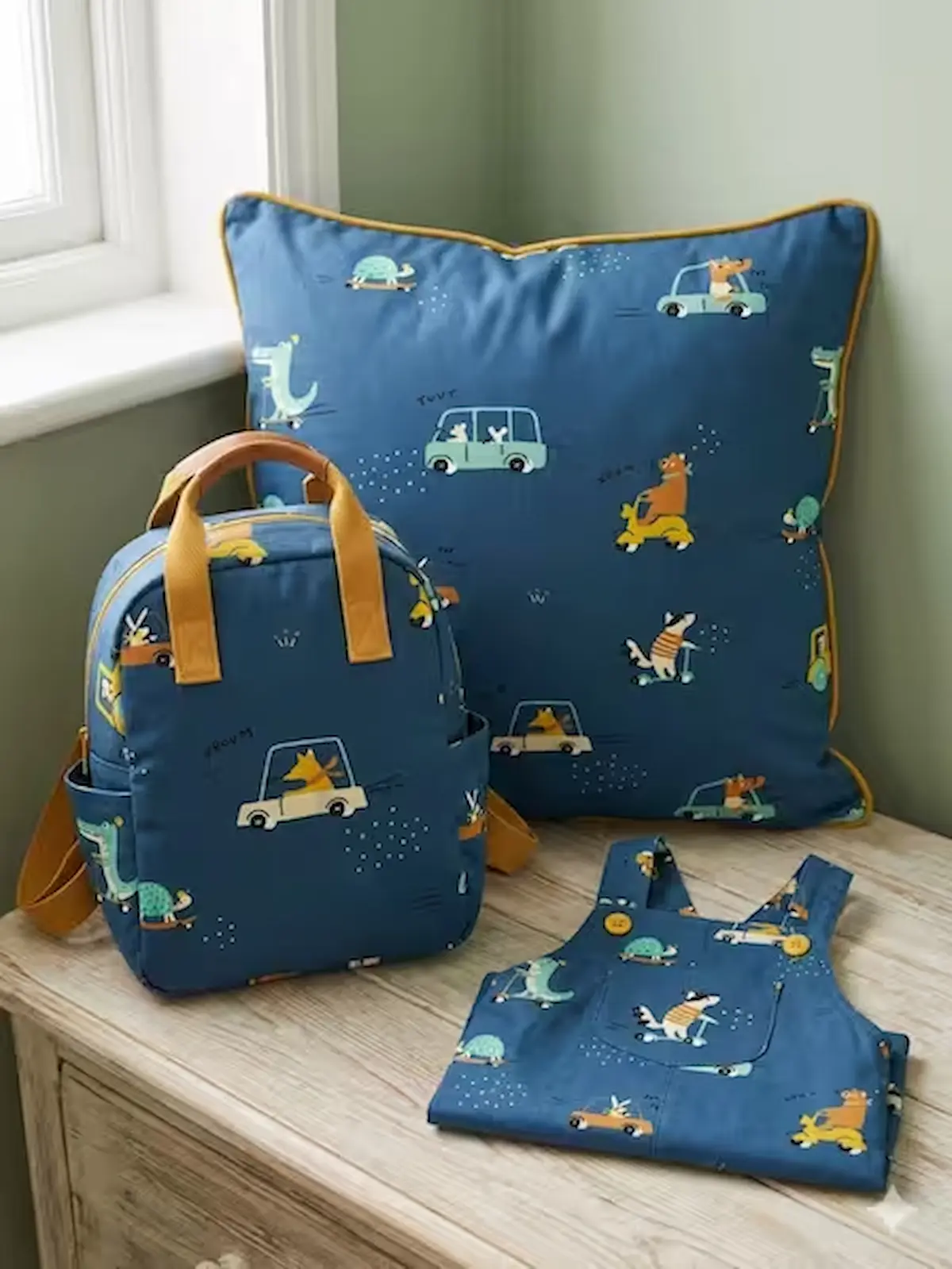 Mittelblauer Baumwollstoff als Webware Meterware mit kindgerechten Tiermotiven in Autos und auf Rollern, präsentiert als genähter Kinderrucksack, Dekokissen und Latzhose.