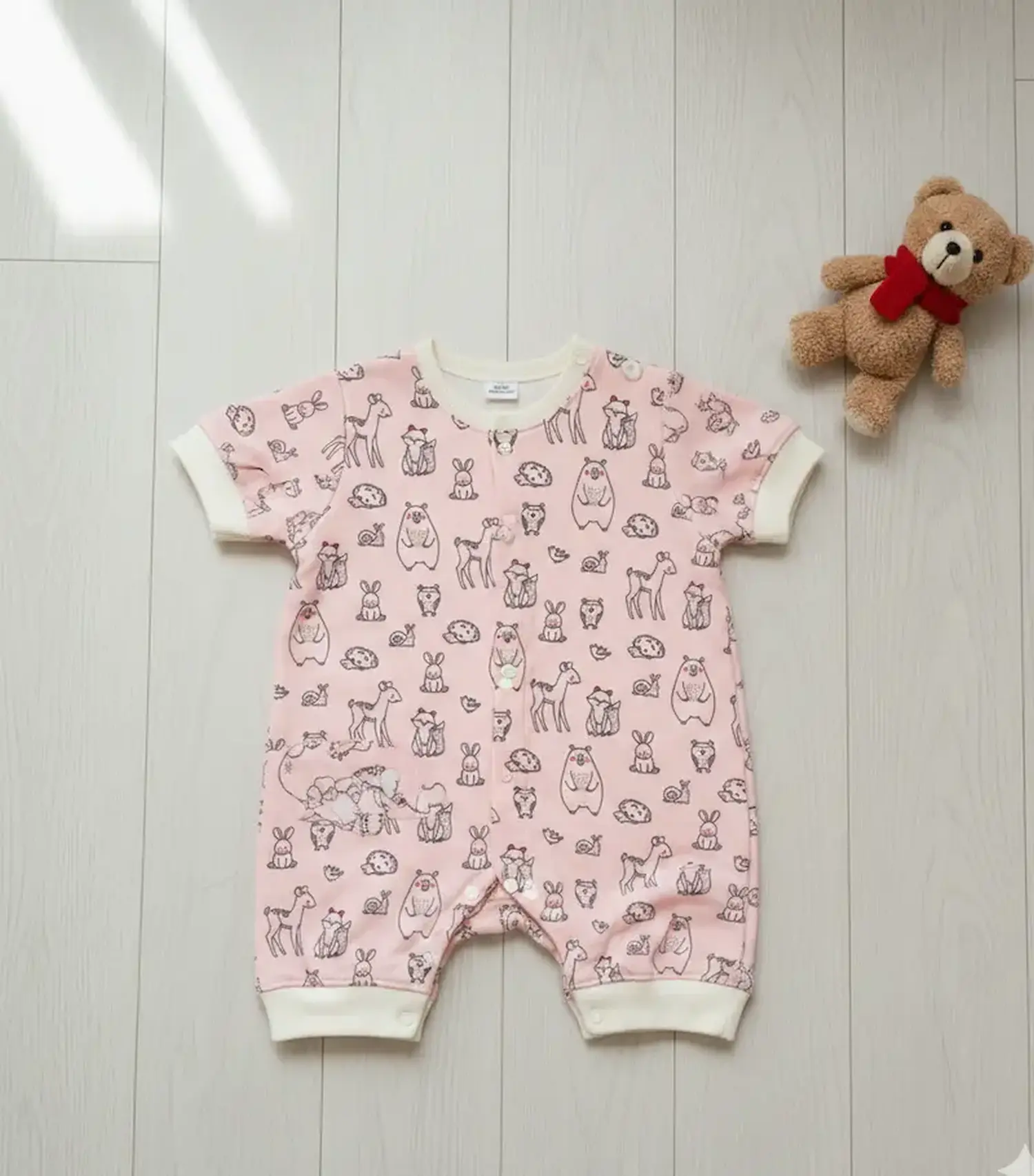 Selbstgenähter Baby-Strampler aus rosa French Terry mit Waldtier-Motiven, kurzen Ärmeln und cremefarbenen Bündchen auf hellem Holzboden mit Teddybär.