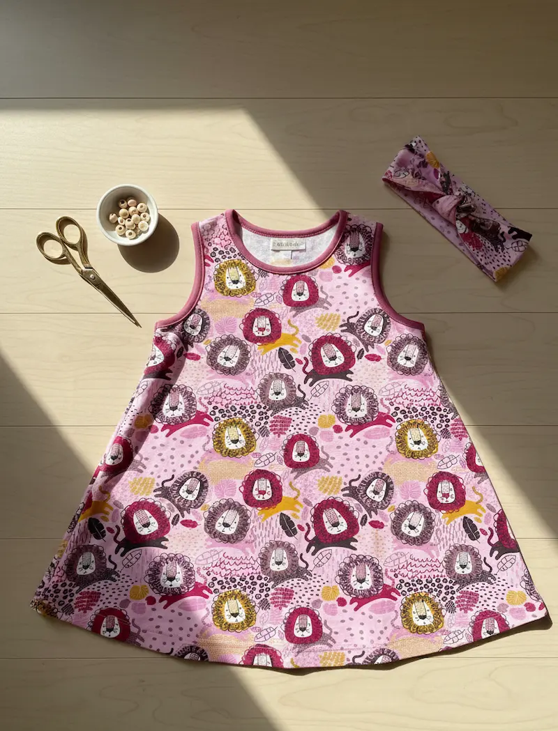 Designbeispiel für French Terry Meterware: Ein selbstgenähtes, ärmelloses Kinderkleid in A-Linie und ein passendes Stirnband aus pinkfarbenem Stoff mit Löwen-Muster liegen flach auf hellem Holzboden neben einer Schere und Holzperlen im Sonnenlicht.