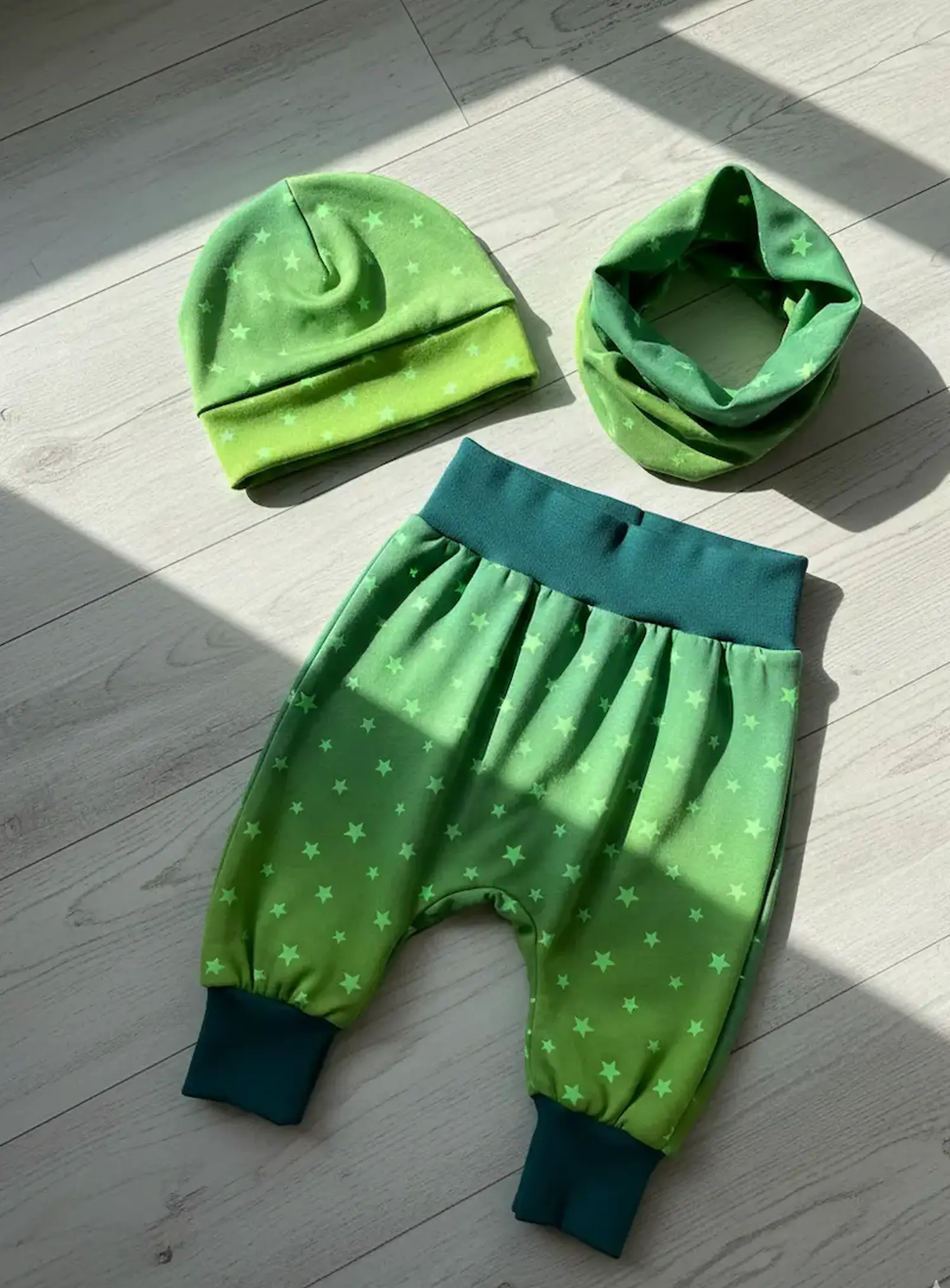 Handgenähtes Baby-Set bestehend aus einer grünen Pumphose, Beanie-Mütze und einem Loop-Schal aus Jerseystoff mit gelbem Sternenmuster und subtilem Farbverlauf, flach liegend auf hellem Holzboden.