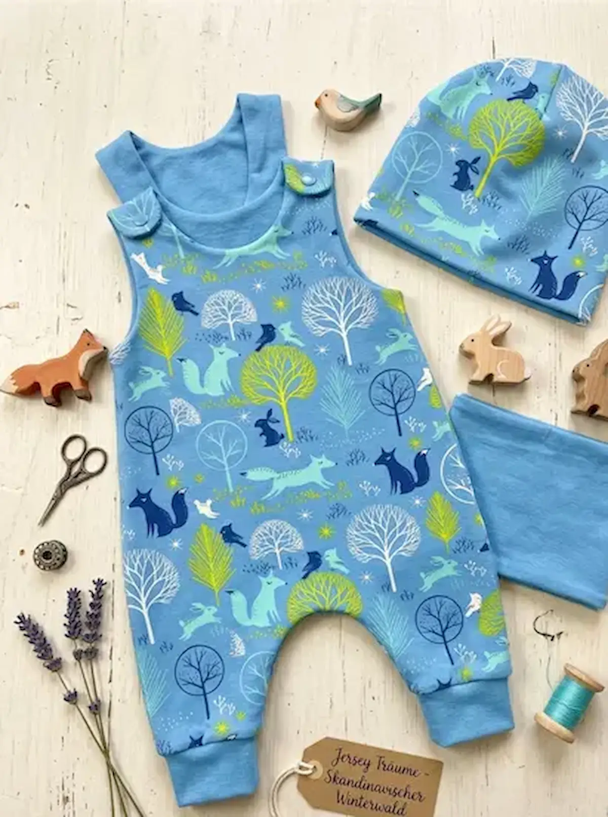 Genähtes Baby-Set aus blauem Baumwolljersey mit Waldtieren: Strampler und Beanie-Mütze mit Fuchs- und Hasen-Motiven auf hellem Holzboden.