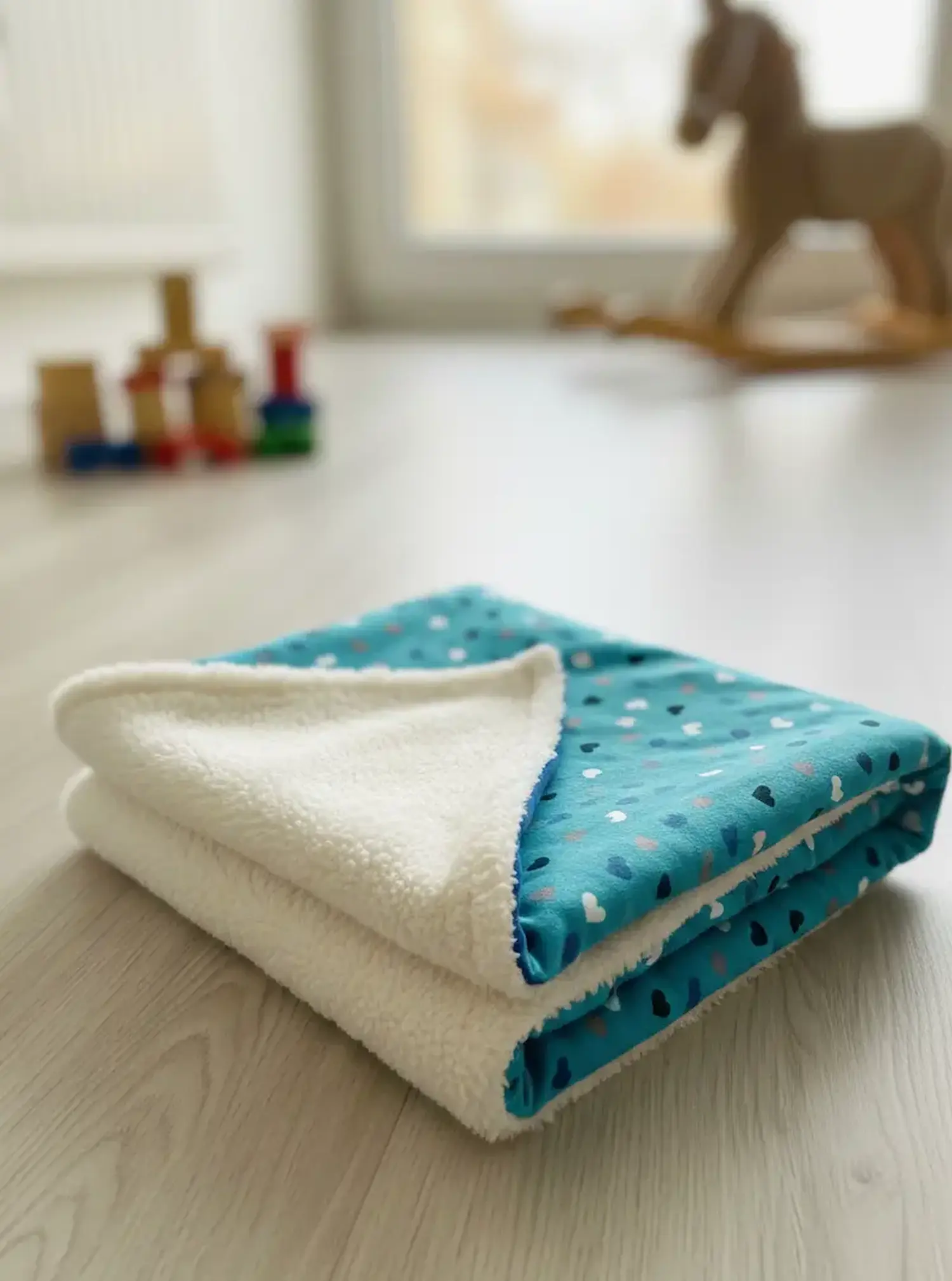 Eine gefaltete Babydecke aus türkisfarbenem Jerseystoff mit kleinen bunten Herzen und weißem, flauschigem Innenfutter liegt auf einem hellen Holzboden; im Hintergrund sind Spielbausteine und ein Schaukelpferd zu sehen.
