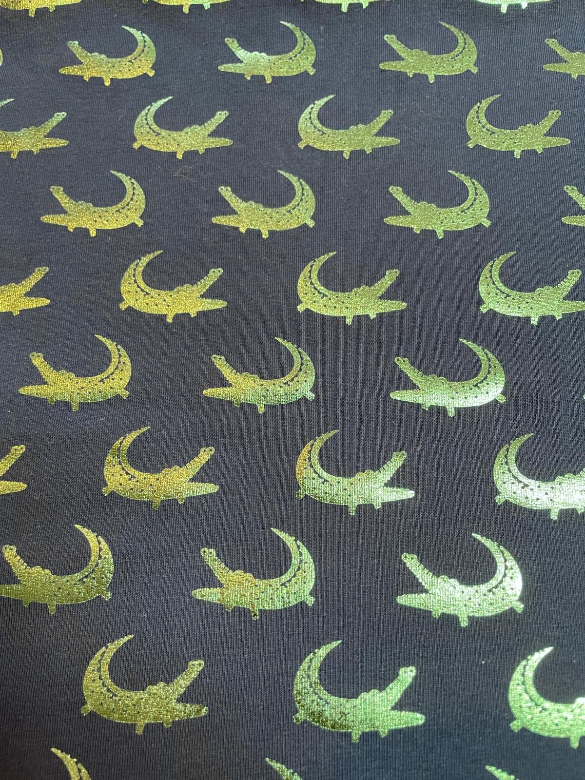 Nahaufnahme von dunkelgrauem Jerseystoff als Meterware, bedruckt mit einem fortlaufenden Muster aus schimmernden, grün-goldenen Metallic-Krokodilen.