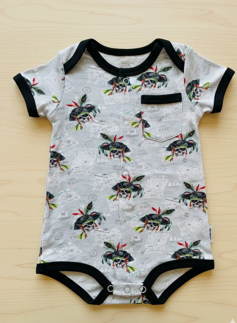 Ein kurzärmeliger Baby-Body mit schwarzer Paspelierung liegt auf einem hellen Holzhintergrund. Der Body ist aus hellgrauem Baumwolljersey gefertigt und mit einem detaillierten Piraten-Motiv (Totenkopf mit Hut und Federn) sowie Schatzkarten-Elementen bedru