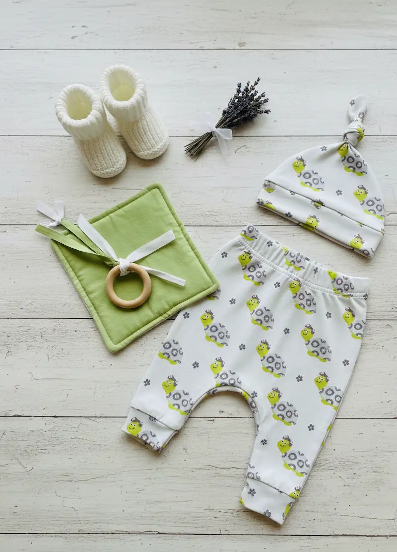 Weicher Jersey-Stoff als Meterware mit niedlichem Schildkröten-Muster in Hellgrau und Grün, präsentiert als genähtes Baby-Set mit Hose und Mütze auf hellem Holzboden.