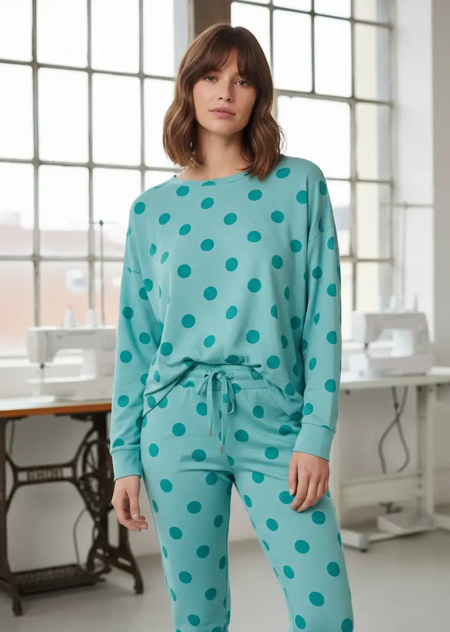 Eine junge Frau in einem hellen Näh-Atelier trägt ein Loungewear-Set bestehend aus Longshirt und passender Hose aus türkisfarbenem Jerseystoff mit großen dunkelblauen Punkten.
