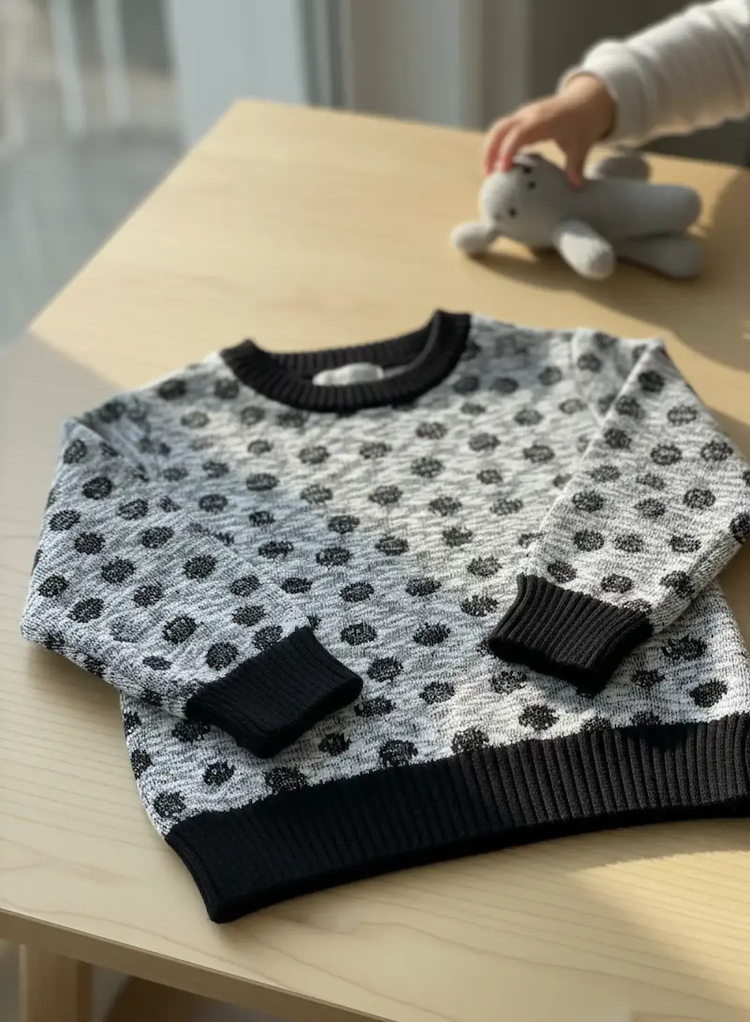 Ein selbstgenähter Pullover aus weichem, grau meliertem Strickstoff mit eingestrickten schwarzen Polka Dots liegt flach auf einem hellen Holztisch. Der Rundhalsausschnitt sowie die Ärmel- und Saumbündchen sind farblich abgesetzt in schwarzem Rippstrick ge