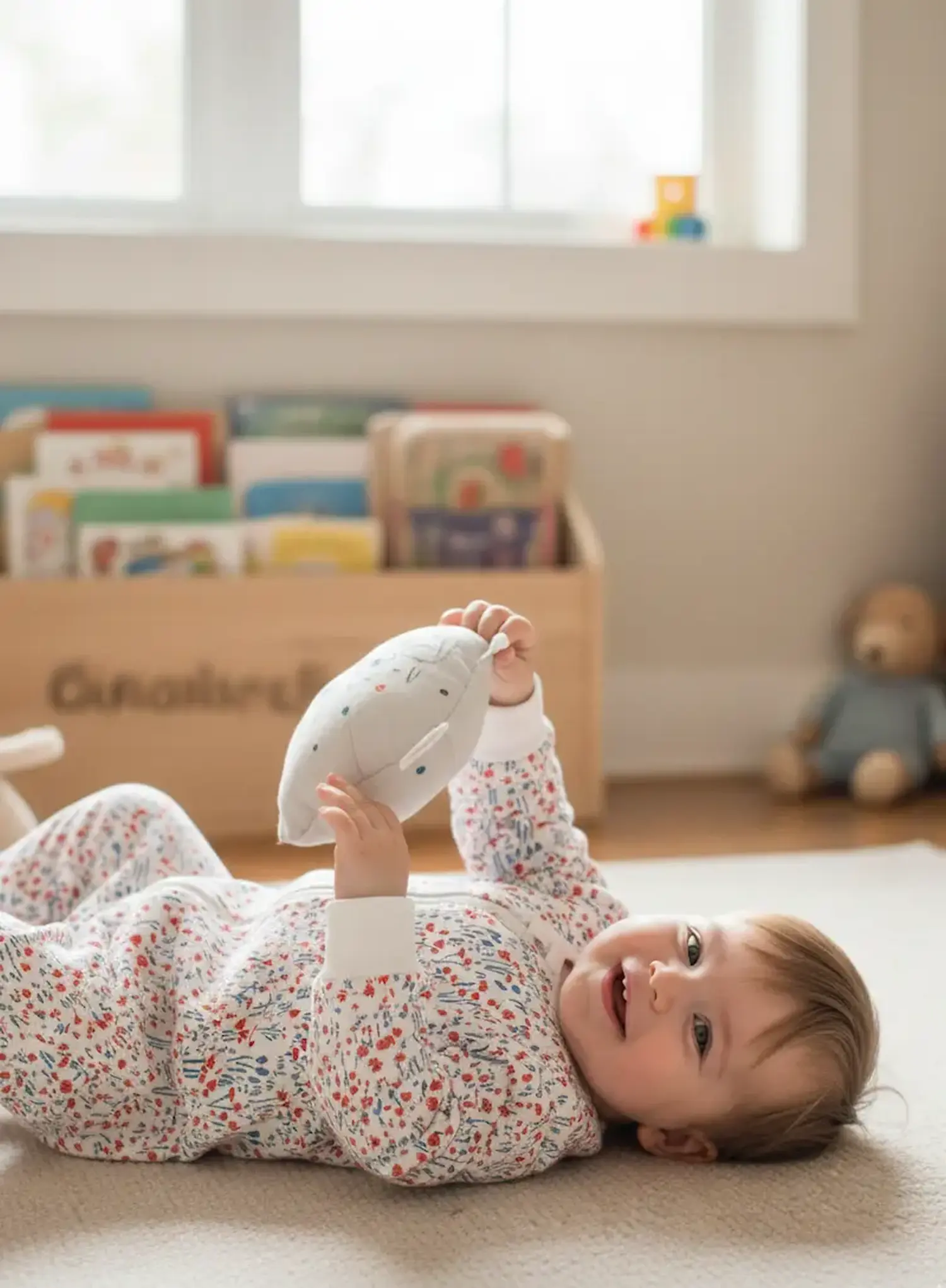Baby trägt einen Strampler aus floralem Sweatstoff mit roten und blauen Blumen. Hochwertige Meterware für Baby- und Kinderkleidung in einer hellen Spielecke.