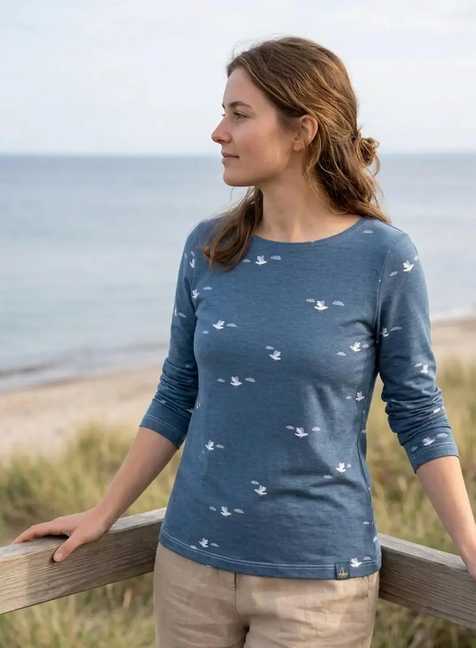 Eine Frau trägt ein blaues Langarmshirt aus meliertem Baumwolljersey mit weißem Möwen- und Wolken-Print an einer Küste.
