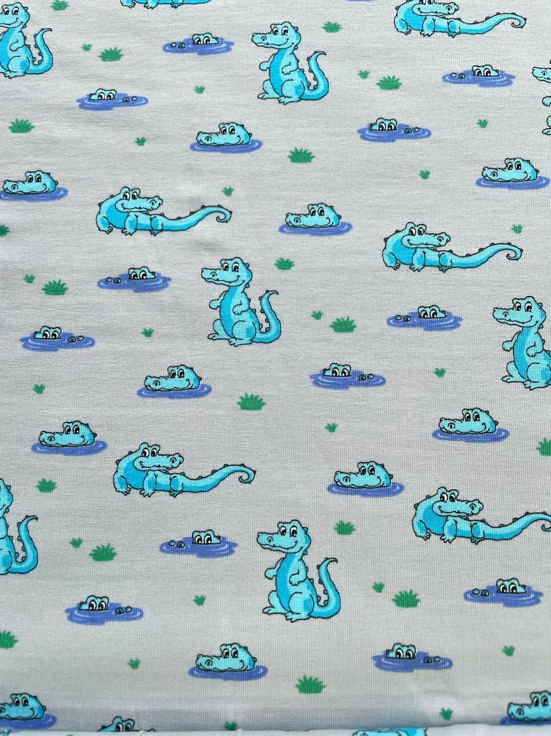 Hochwertiger Baumwolljersey als Meterware in Hellgrau Melange, bedruckt mit einem niedlichen Comic-Muster aus hellblauen Krokodilen. Die Alligatoren sitzen auf dem Land, schwimmen im Wasser oder gucken aus kleinen Sumpflöchern hervor, verziert mit kleinen