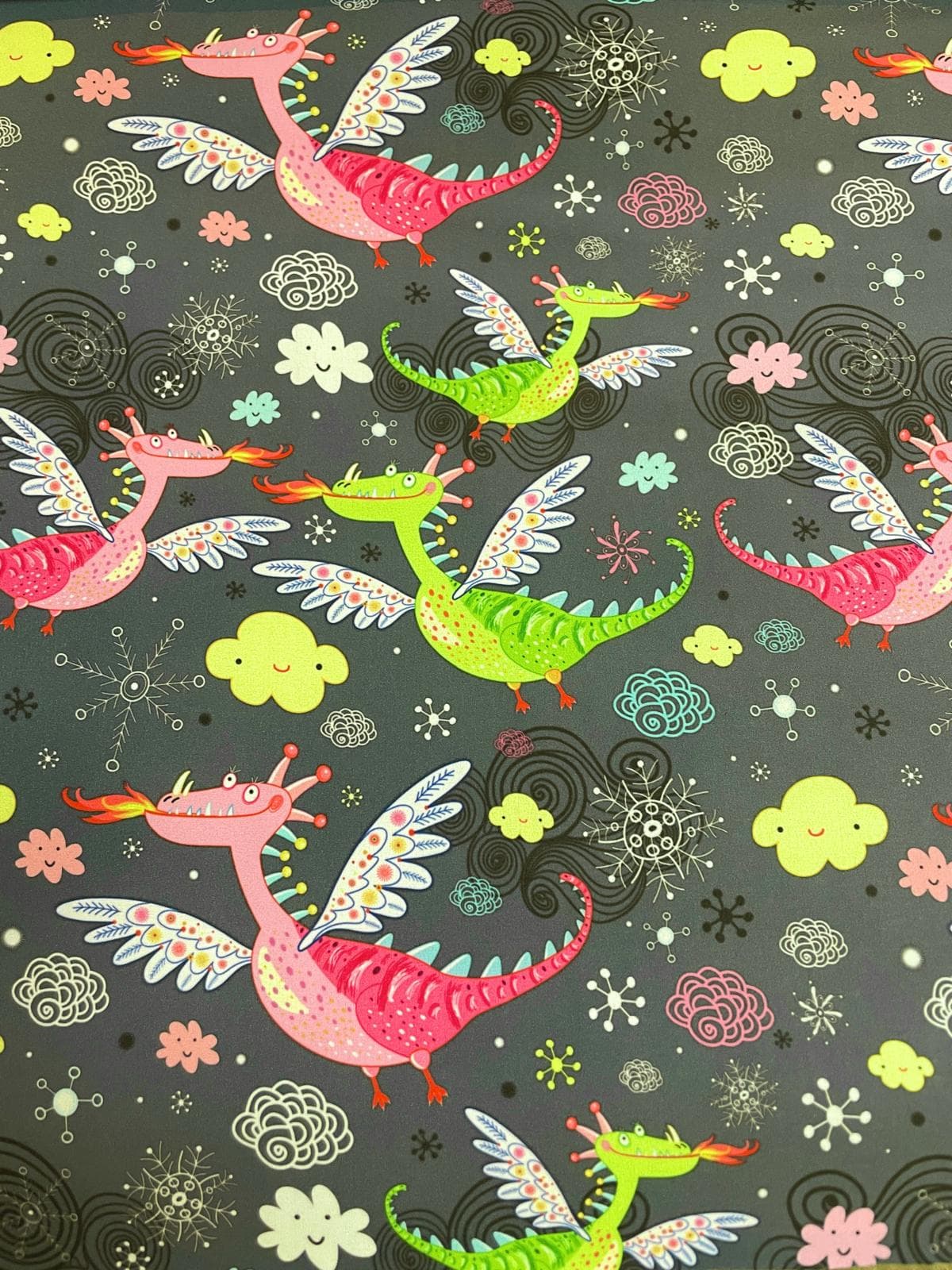 Hochwertiger Softshell-Stoff mit Drachen-Motiv auf dunkelgrauem Hintergrund. Zu sehen sind freundliche, feuerspeiende Drachen in Pink und Grün mit bunt verzierten Flügeln. Das Muster wird ergänzt durch kleine lachende Wolken, Schneeflocken und Kringel-Ele