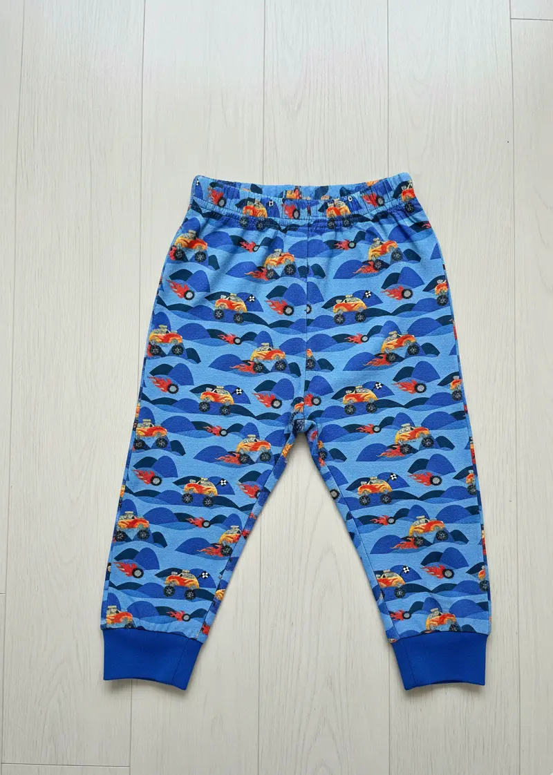 Eine flach liegende Kinderhose aus blauem Baumwolljersey mit einem Allover-Print von Monster-Trucks mit Flammenmotiven vor einer hügeligen blauen Landschaft. Die Hose ist mit passenden royalblauen Bündchen an den Beinen und einem elastischen Bund versehen