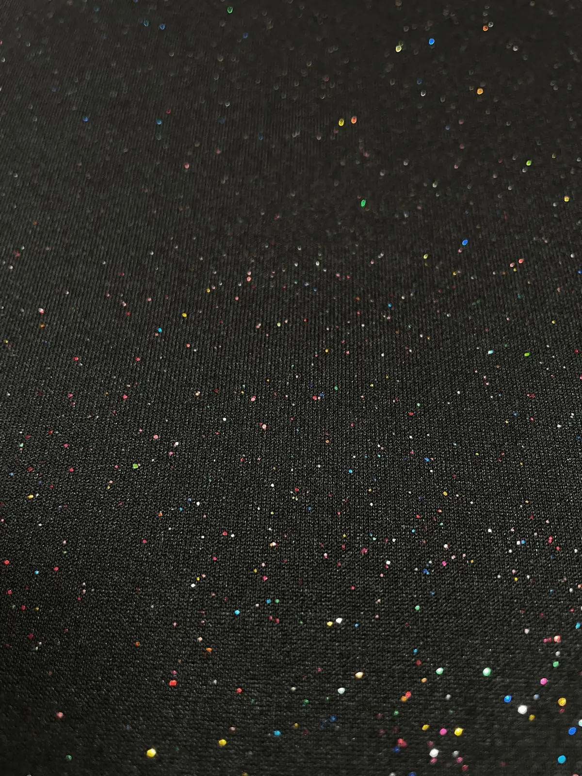 Nahaufnahme eines schwarzen Jerseystoffs, der mit vielen kleinen, bunten Glitzerpunkten (Multicolor) übersät ist und im Licht funkelt.