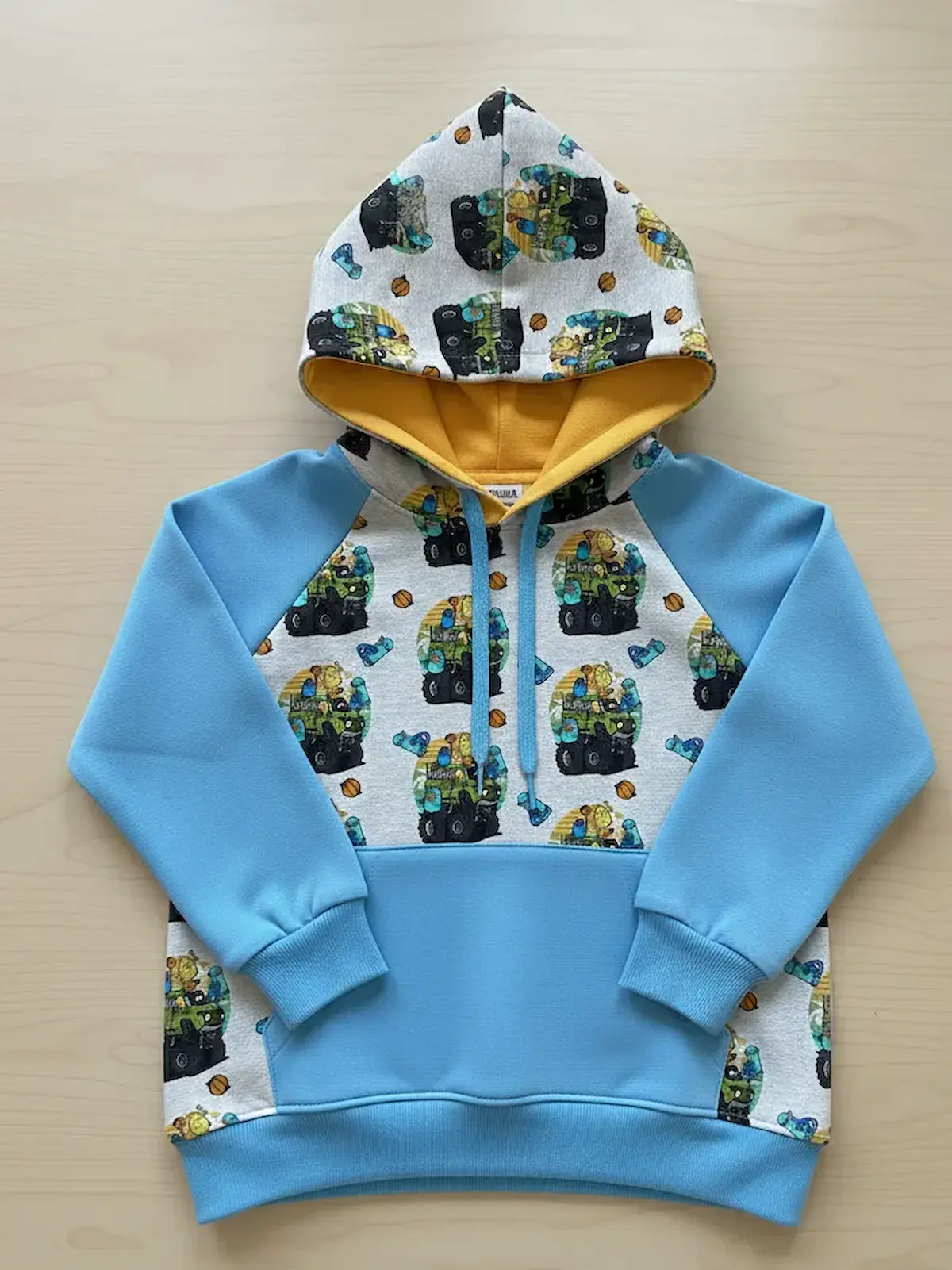 Genähter Kinder-Raglan-Hoodie aus grauem Monster-Truck-French-Terry kombiniert mit hellblauen Ärmeln und senfgelbem Kapuzenfutter.