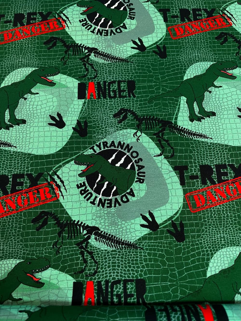 Ein grüner Sommersweat-Stoff für Kinder mit einem detailreichen Dinosaurier-Druck. Das Muster zeigt T-Rex-Illustrationen, Skelette, Fußabdrücke und Warnschilder wie 'Danger' und 'Tyrannosaurus Adventure'. Der Hintergrund hat eine grüne Krokodil-Haut-Optik