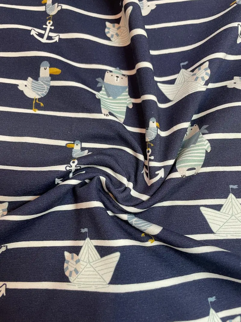 Ein kunstvoll drapierter Baumwolljersey-Stoff als Meterware mit dunkelblauen und weißen Querstreifen. Durch den weichen Faltenwurf kommen die kleinen, gedruckten Motive wie Bären im Matrosenlook, Möwen und Papierschiffchen schön zur Geltung.