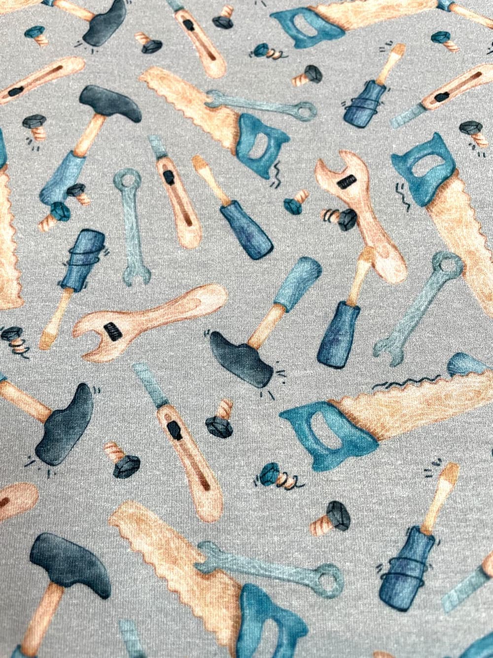 Flach liegender, hellgrau melierter Jerseystoff als Meterware, vollflächig bedruckt mit bunten, illustrierten Werkzeugen wie Sägen, Hämmern, Schraubenschlüsseln und Schrauben.