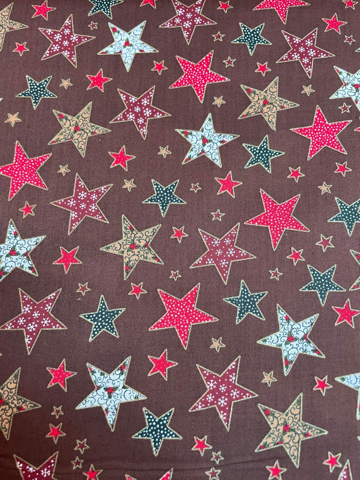 Festlicher Baumwollstoff als Meterware mit bunt gemusterten Sternen in Rot, Grün, Hellblau und Beige mit Ornamenten und Schneeflocken auf braunem Hintergrund.
