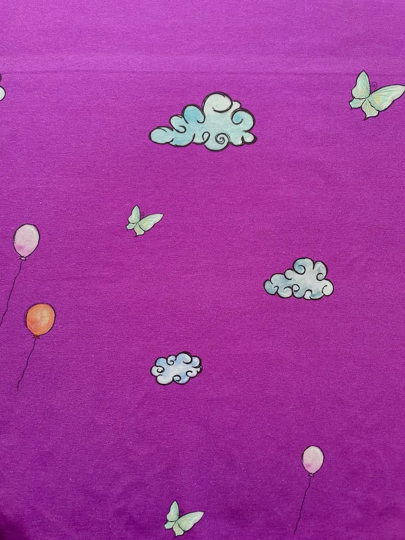 Nahaufnahme eines lilafarbenen Baumwolljersey-Stoffes mit einem verspielten Panel-Design. Das Motiv zeigt hellblaue, illustrierte Wolken, zarte hellgrüne Schmetterlinge und schwebende Luftballons in Rosa und Orange.
