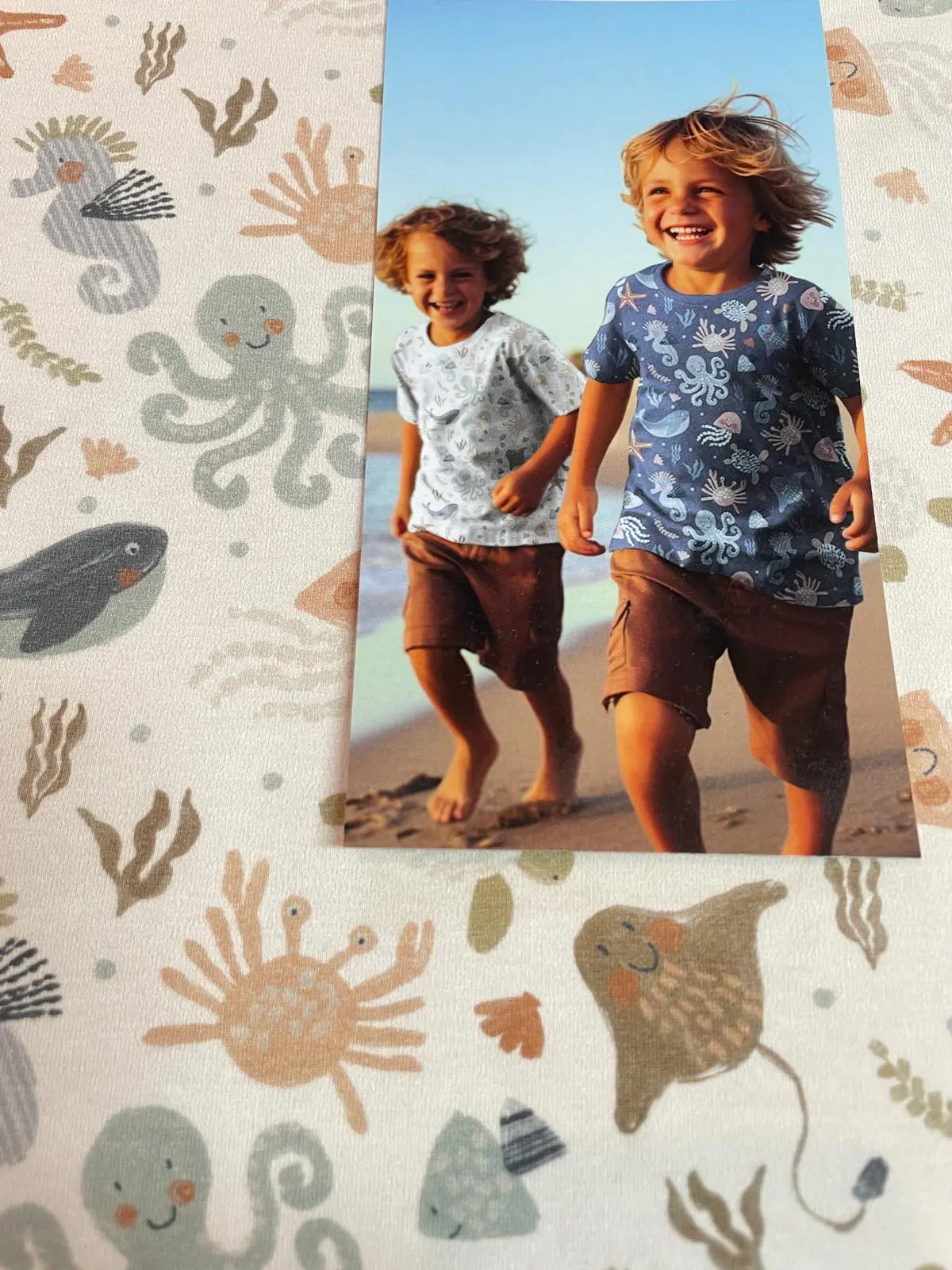 Ein gedrucktes Foto liegt auf einem cremeweißen Ozean-Stoff und zeigt zwei fröhliche, am Strand entlanglaufende Kinder, die selbstgenähte T-Shirts aus dem hellen und dunkelblauen Meerestiere-Jerseystoff tragen.