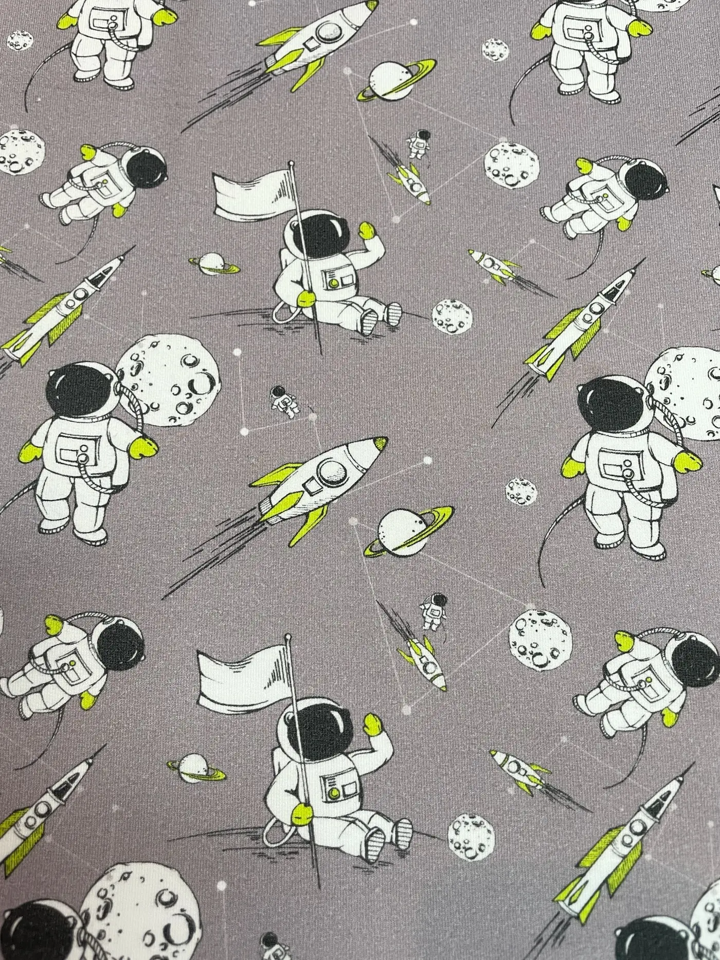 Hochwertiger French Terry Sommersweat in hellem Grau. Das Muster zeigt detaillierte Illustrationen von Astronauten mit Flaggen, startenden Raketen, Planeten mit Ringen und Monden. Markante Farbakzente in Neon-Gelb/Lime ergänzen das schwarz-weiße Weltraum-