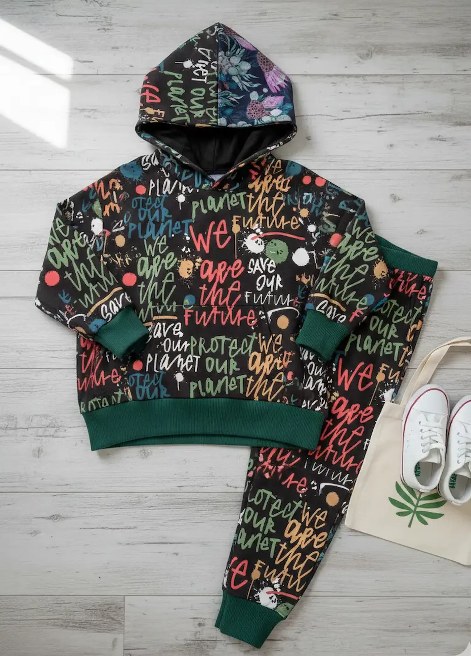 Zweiteiliges Kinder-Outfit aus schwarzem French Terry mit bunten 'Save our Planet' Graffiti-Schriftzügen, Kapuzenpullover und Hose mit dunkelgrünen Bündchen auf hellem Holzboden