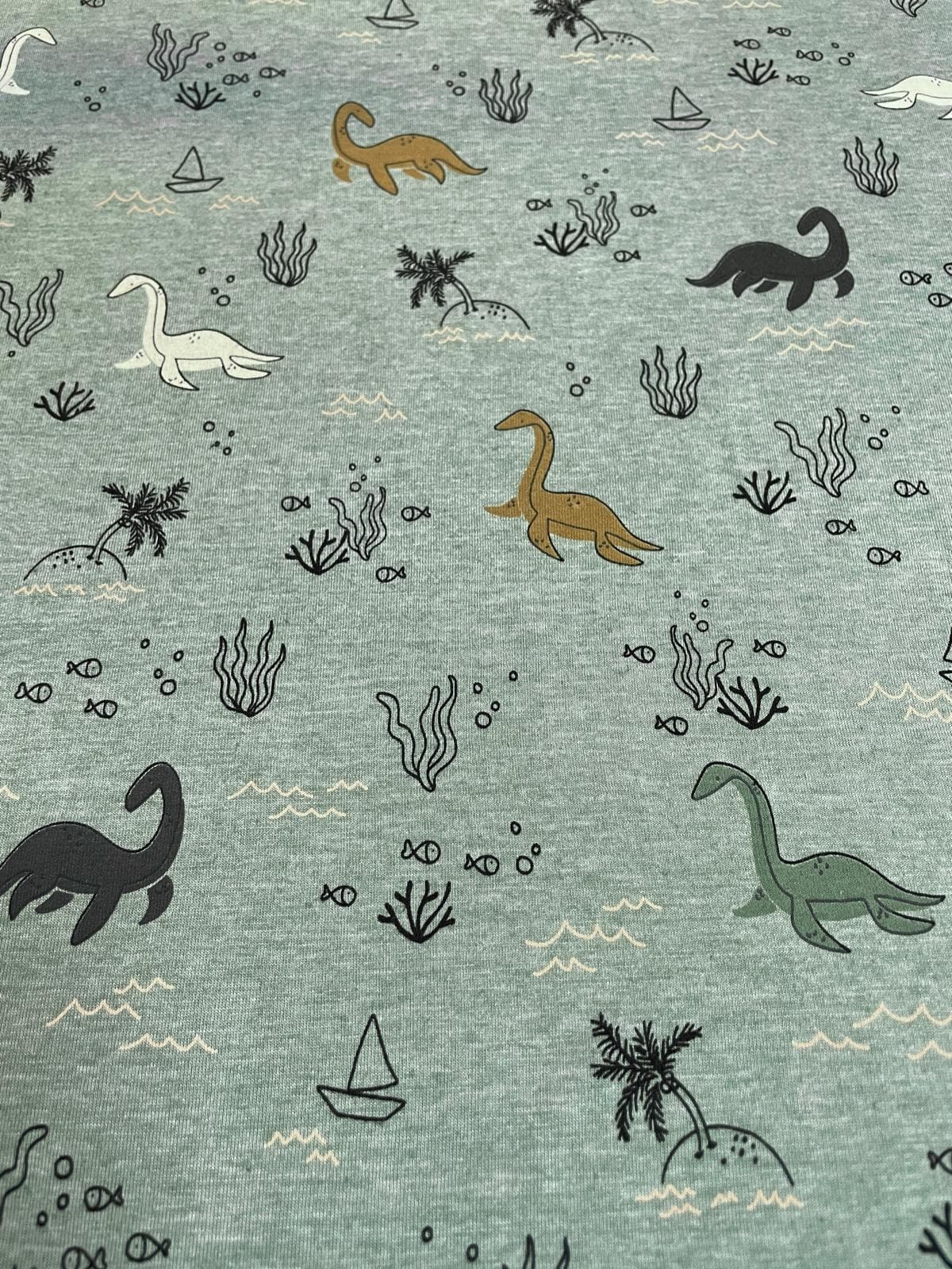 Salbeigrün melierter Baumwolljersey-Stoff als Meterware, flach fotografiert und bedruckt mit einem verspielten Muster aus verschiedenen Langhals-Dinosauriern im Wasser, kleinen Palmeninseln, Fischen, Algen und Segelbooten.