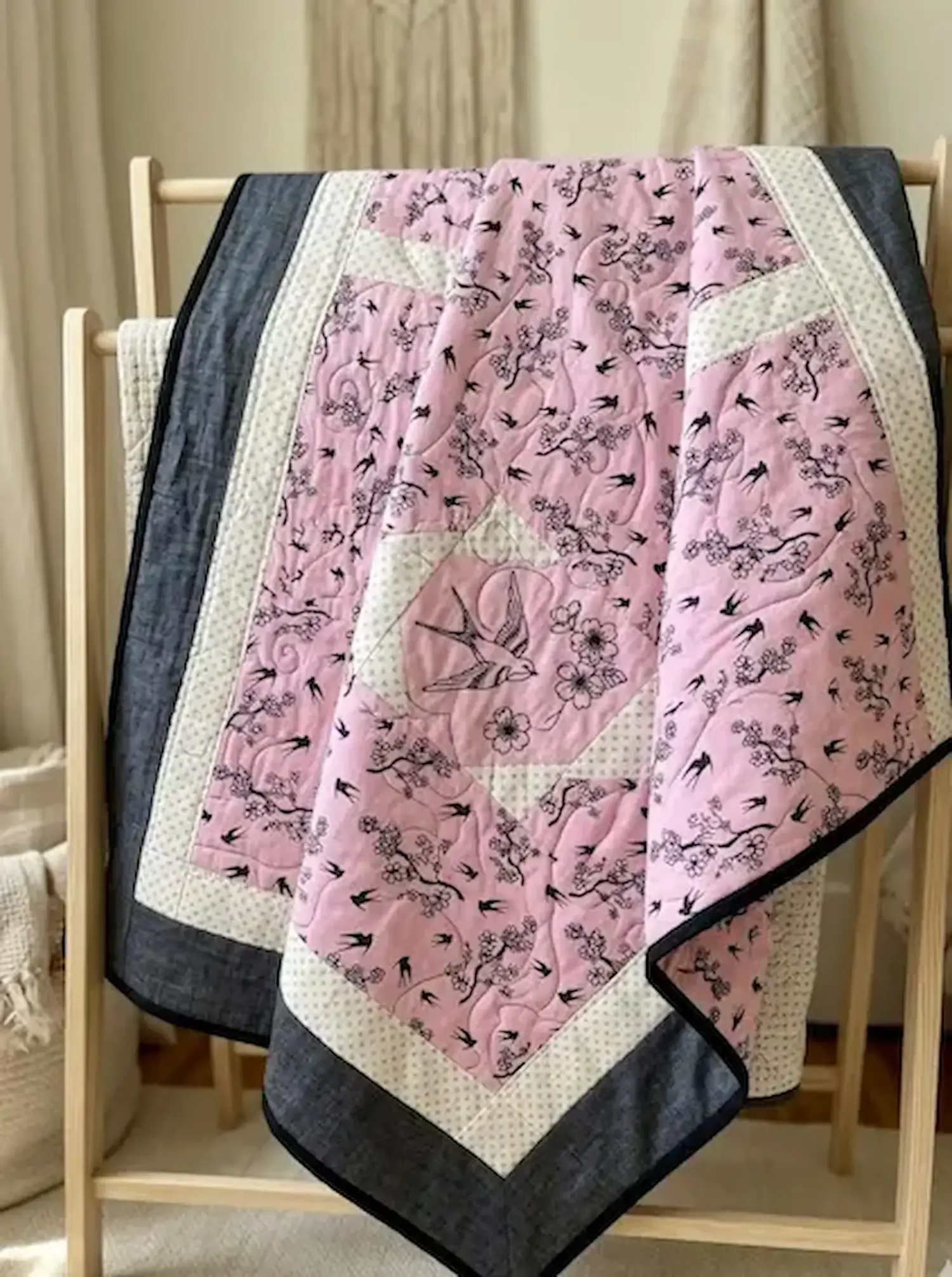 Handgefertigte Babydecke aus rosa Baumwollstoff mit schwarzen Schwalben- und Blumenmotiven auf einem Holzständer.