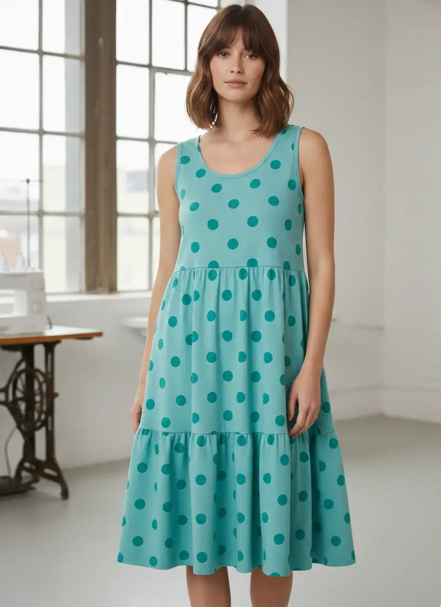 Eine junge Frau steht in einem hellen Näh-Atelier vor einer Nähmaschine und trägt ein sommerliches Stufenkleid aus türkisfarbenem Jerseystoff mit großen dunkelblauen Punkten.