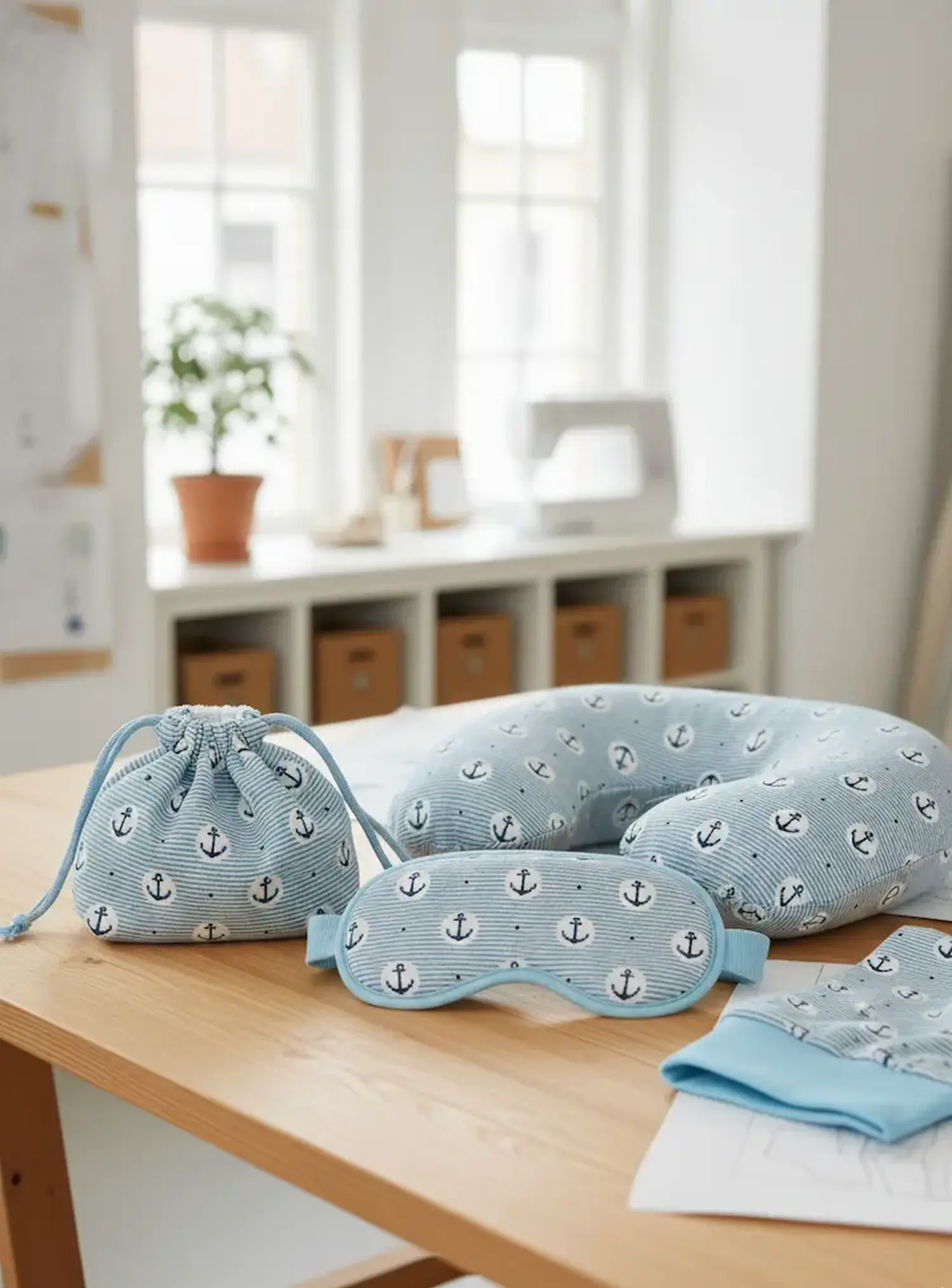 Ein handgenähtes Set bestehend aus einem Tunnelzugbeutel, einem Nackenkissen und einer Schlafmaske aus hellblau gestreiftem Jerseystoff mit Ankermotiv auf einem Holztisch.