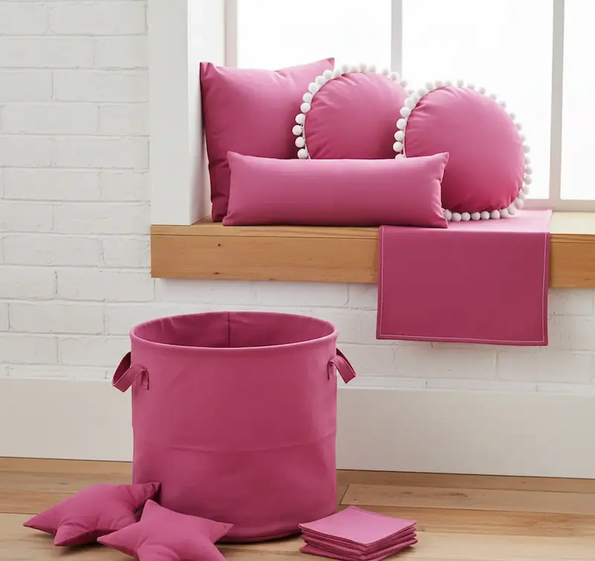 Farbenfrohe Zimmer-Dekoration aus magentafarbener Baumwoll-Popeline, bestehend aus einem großen runden Stoffkorb auf dem Holzboden, Sternenkissen, einem Tischläufer und Dekokissen mit weißer Pompon-Borte auf einer Fensterbank.