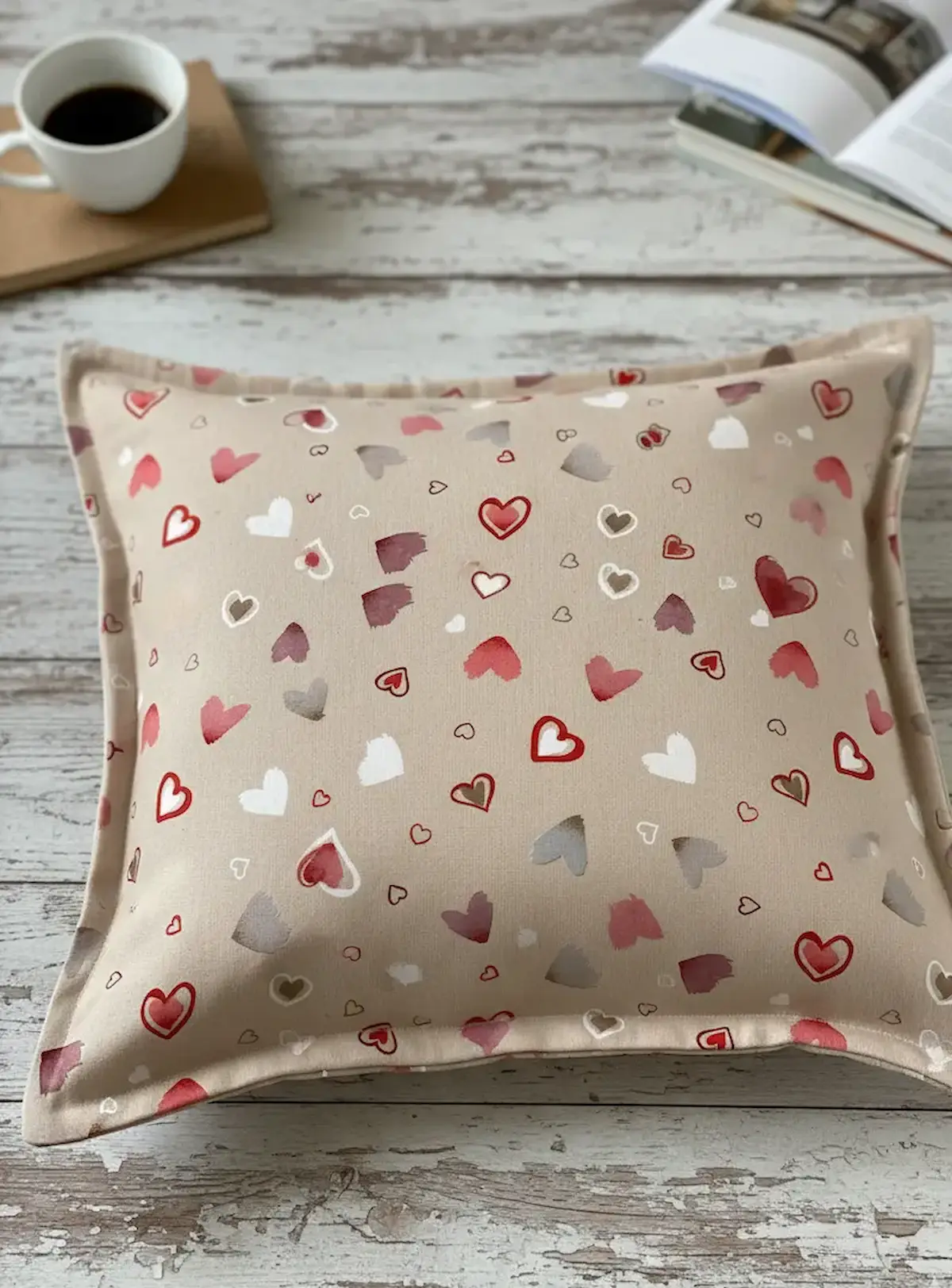 Quadratisches Zierkissen mit Stehsaum aus beigem Herz-Dekostoff, platziert auf einem rustikalen Holztisch neben einer Kaffeetasse.