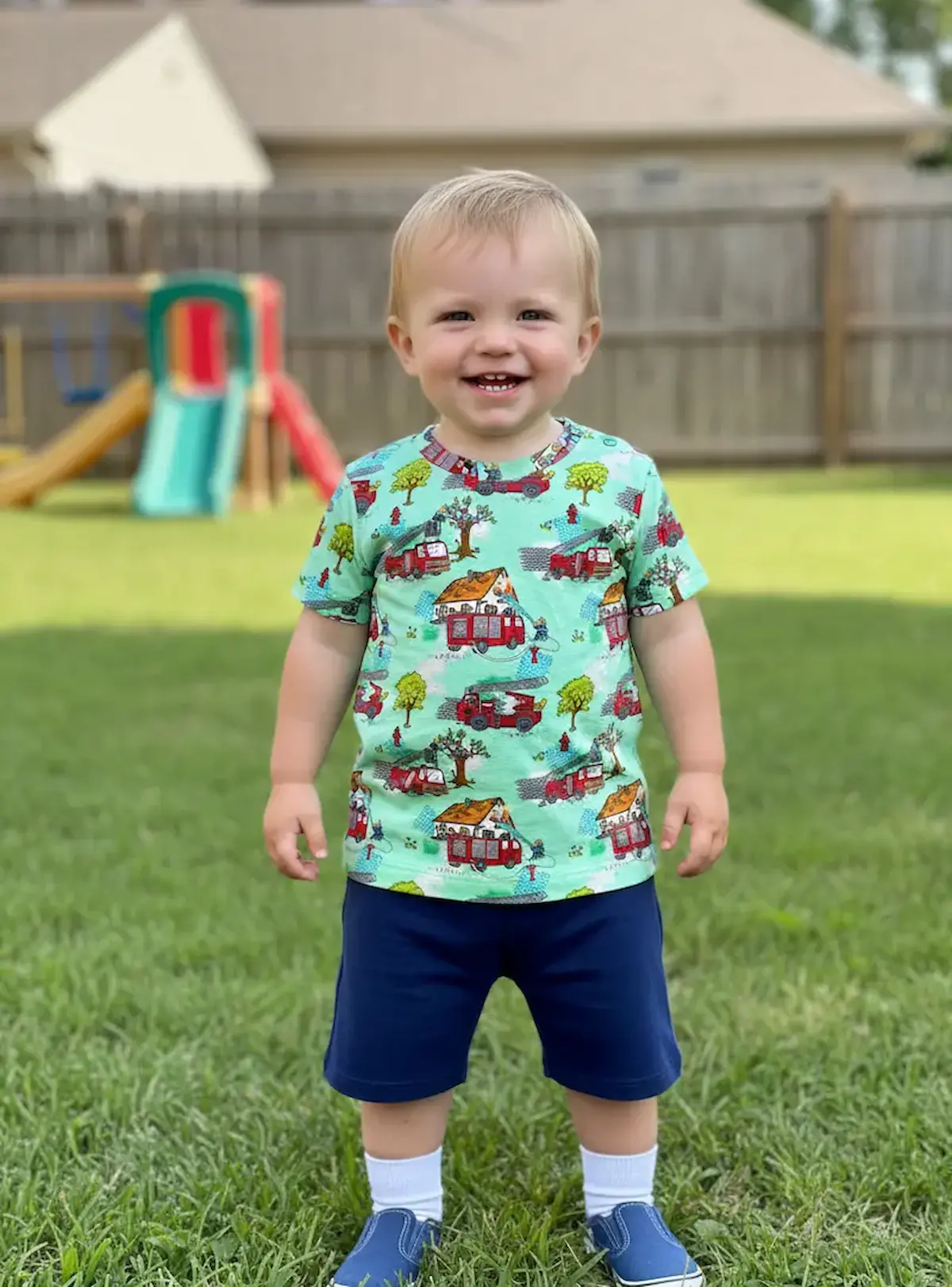 Kleinkind im Garten trägt ein selbstgenähtes T-Shirt aus hellgrünem Jersey-Stoff mit bunten Feuerwehrauto-Motiven.