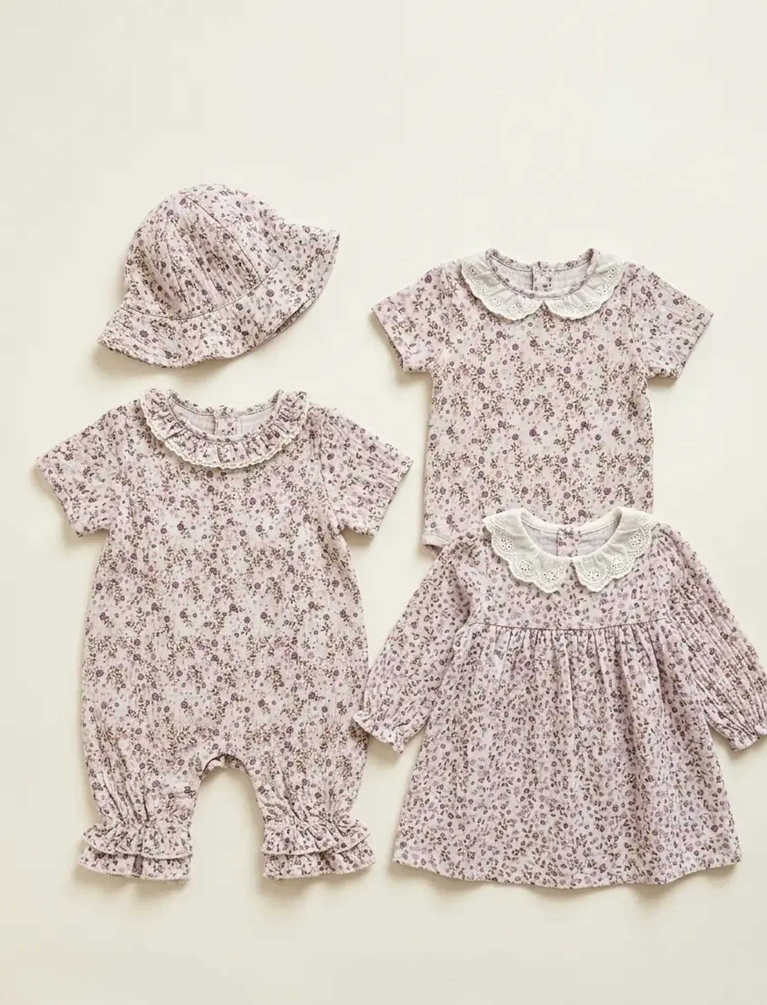 Flachgelegte Ansicht von Babykleidung aus fliederfarbenem Musselin-Stoff mit feinem Blumenmuster, bestehend aus einem Strampler mit Rüschen, einem Kleidchen mit Spitzenkragen, einem Shirt und einem passenden Sonnenhut.
