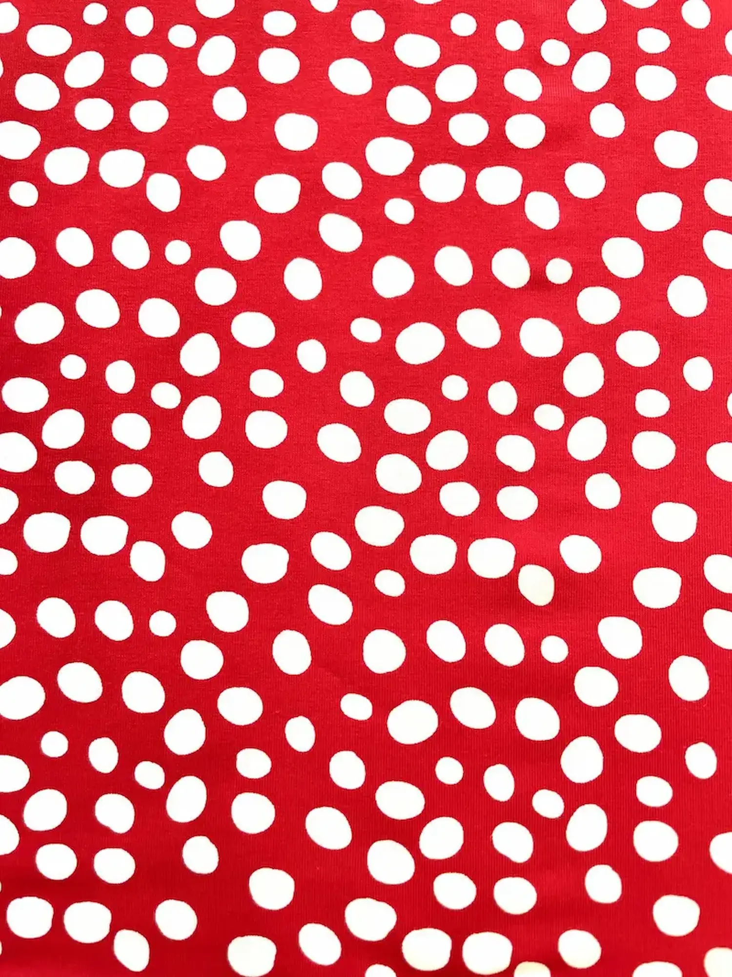 Draufsicht auf einen leuchtend roten Baumwolljerseystoff mit unregelmäßigen weißen Punkten (Polka Dots) als elastische Meterware für Nähprojekte.