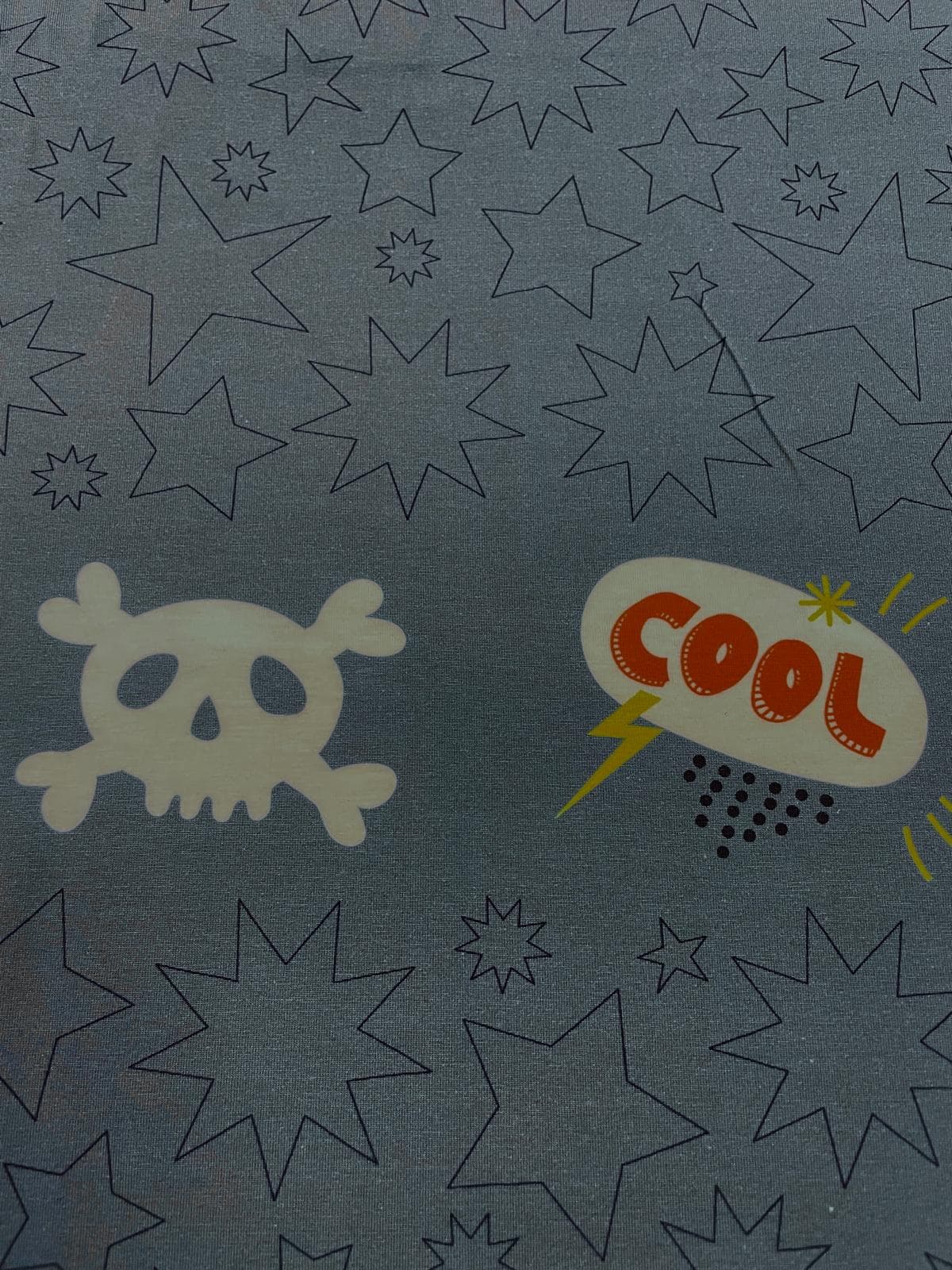 Nahaufnahme eines hochwertigen Baumwolljersey-Panels in Blaugrau. Zu sehen sind ein großer, cremeweißer Comic-Totenkopf mit gekreuzten Knochen sowie eine orangefarbene „COOL“-Sprechblase mit gelbem Blitz und schwarzen Punkten. Der Hintergrund ist mit fein