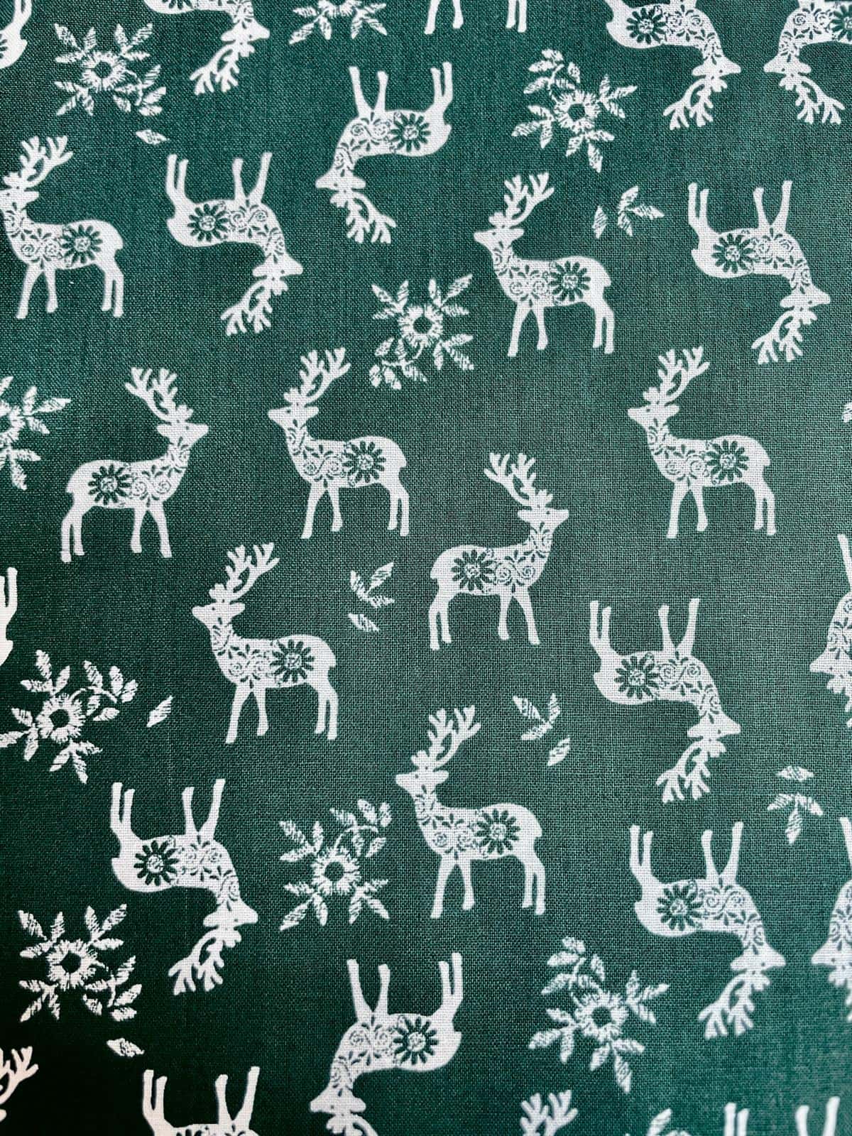 Eleganter Baumwollstoff als Meterware mit stilisierten Hirschen, Rehen mit floralen Mustern und Schneeflocken in Weiß auf dunklem Tannengrün.