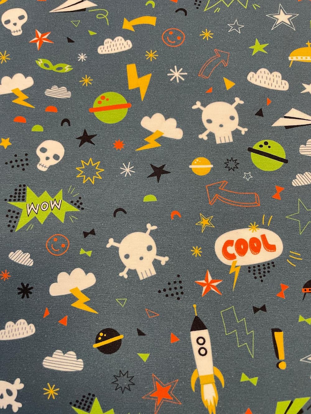 Hochwertiger Baumwolljersey als Meterware in Blaugrau / Jeansblau mit einem bunten Allover-Comic-Muster. Das Streumuster besteht aus kleinen, fliegenden Weltraum-Raketen, weißen Totenköpfen, Blitzen, Wolken, Sternen, Planeten und Sprechblasen mit den Wört
