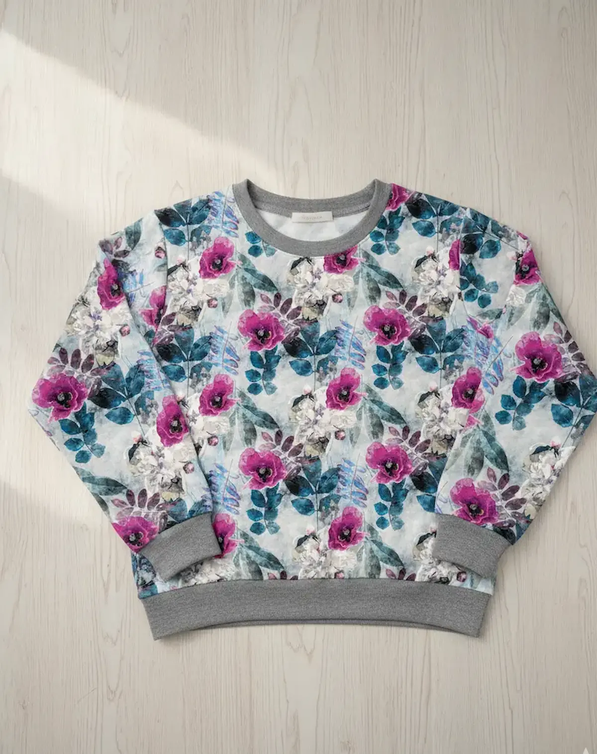 Genähtes Damen-Sweatshirt aus French Terry mit Mohnblumen-Aquarell-Print und grauen Bündchen auf hellem Holzhintergrund.