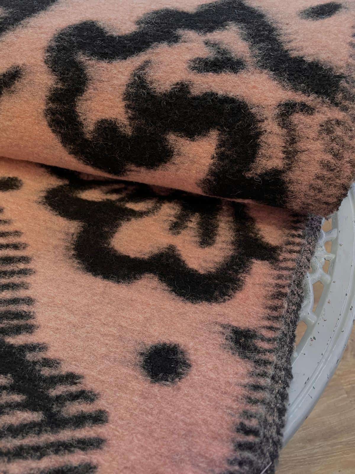 Hochwertiger, dicker Wollstoff als Meterware in einem sanften Altrosa / Apricot, gemustert mit großen, flauschigen Ornamenten im schwarzen Ikat-Stil. Die Nahaufnahme zeigt die weiche, leicht angeraute Textur des dicht gewebten Stoffes, der elegant gefalte