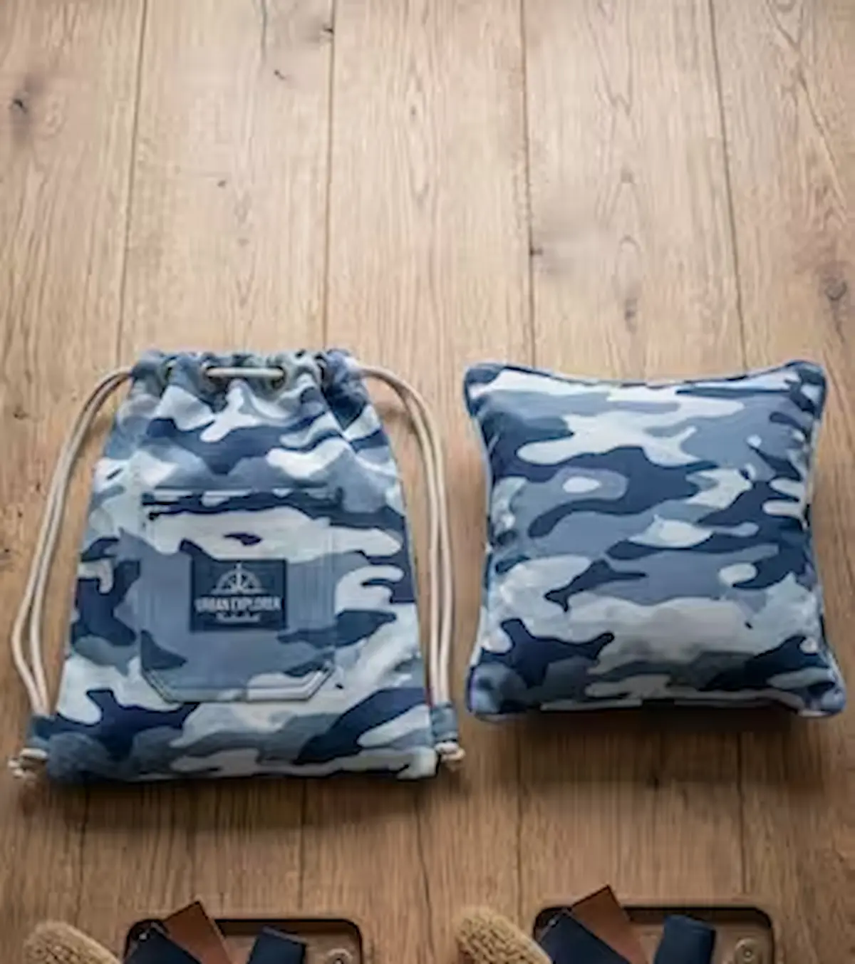 Robuster Baumwoll-Popeline-Stoff als Meterware mit Camouflage-Muster in Blau-, Grau- und Weißtönen, präsentiert als genähter Rucksack und Kissen auf einem Holzfußboden.